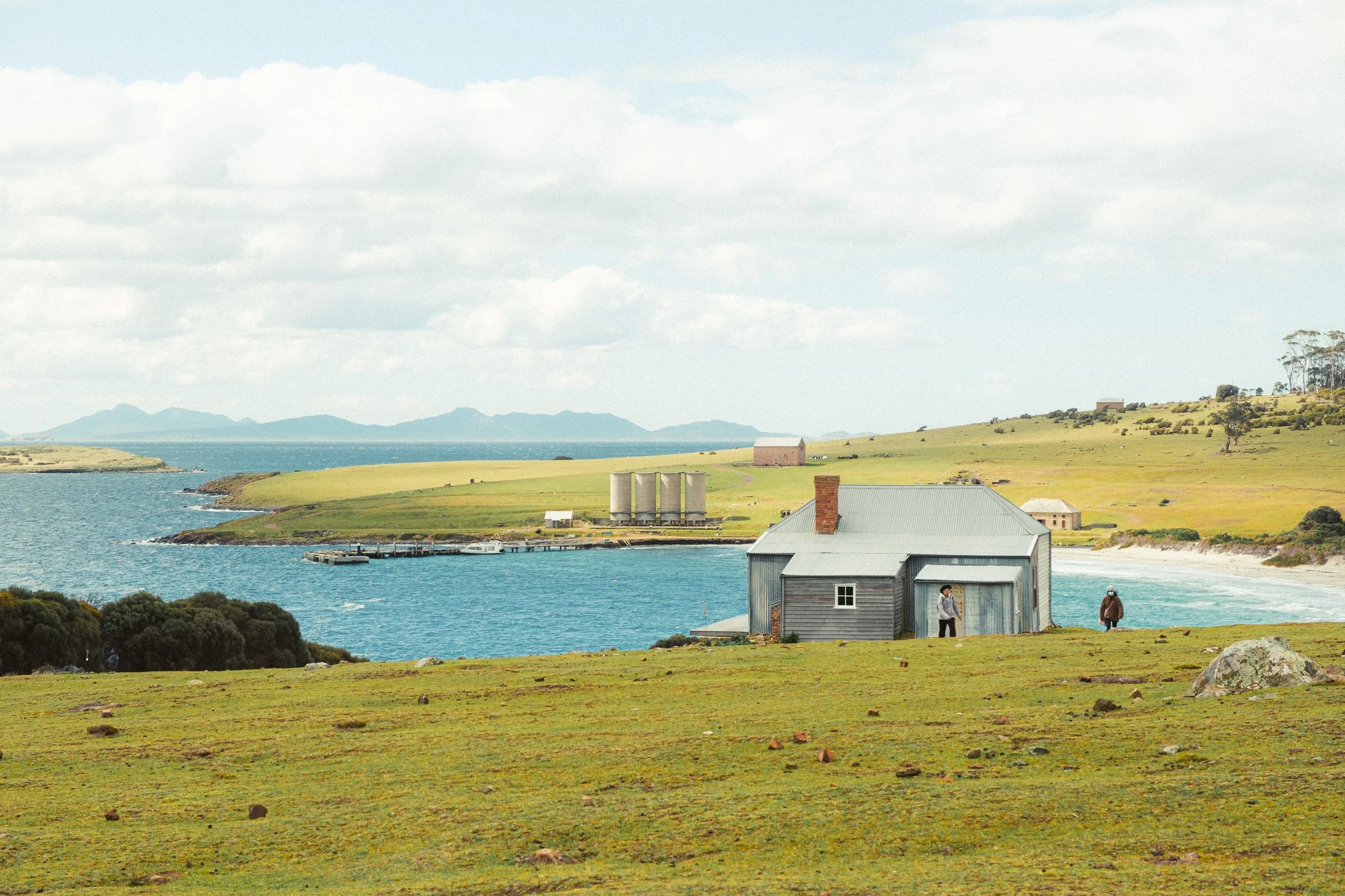 Coastal landscape with grassy foreground, houses, and a body of water, mountains in the distance, and a partly cloudy sky.