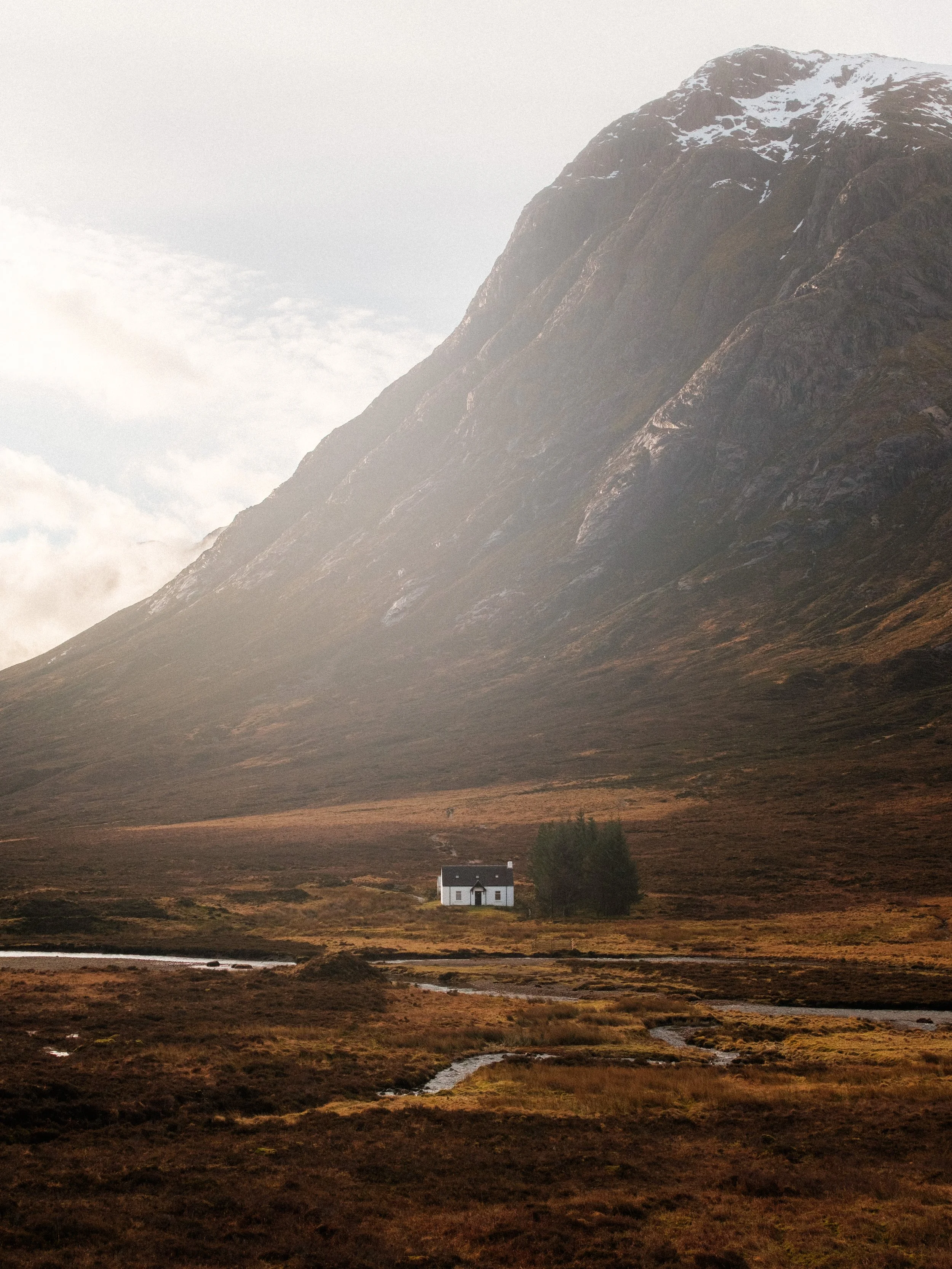 A small white house with a dark roof sits in a wide open field in front of a large mountain with patches of snow.