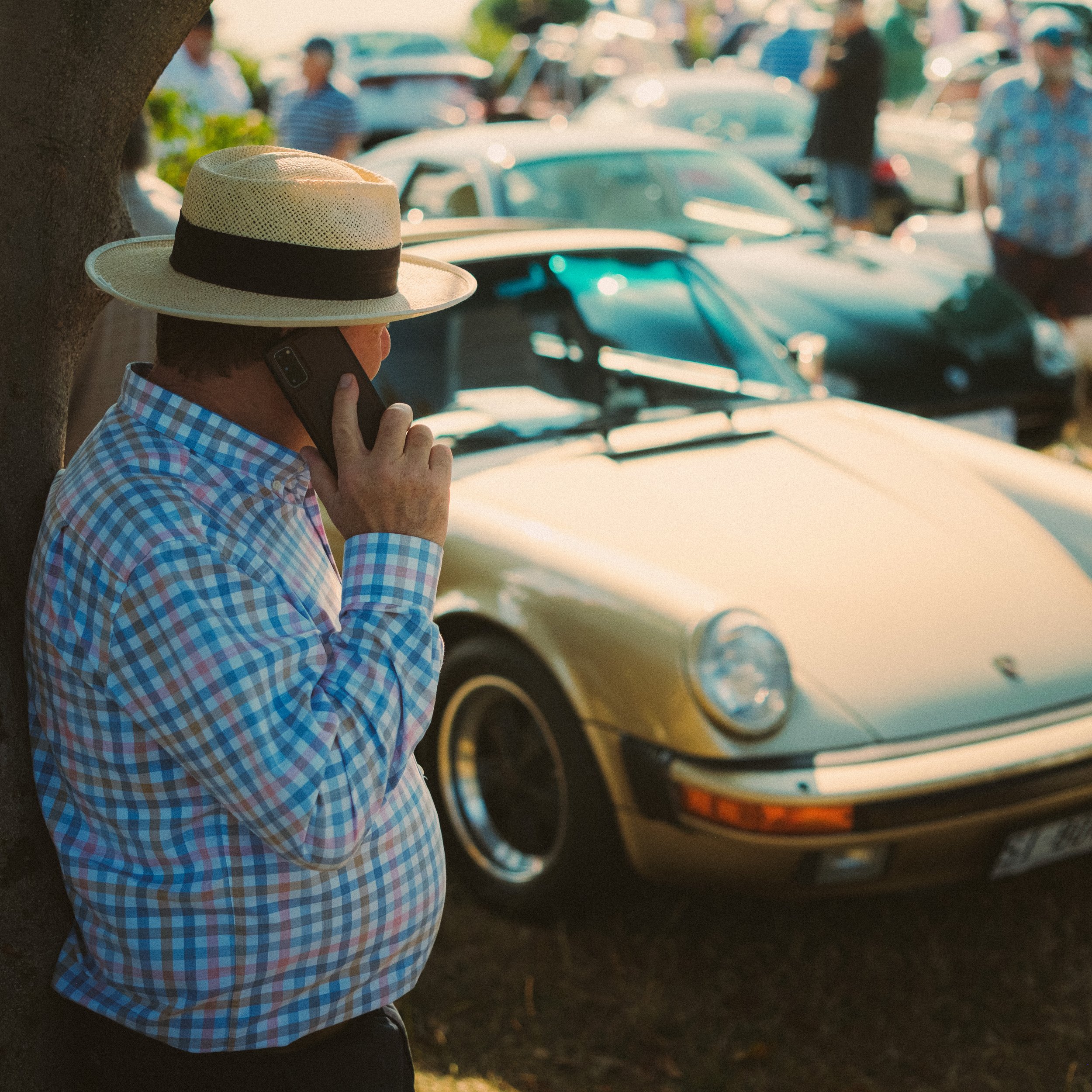 A man wearing a checked shirt and a straw hat is talking on his cell phone, leaning against a tree. In the background, there are several classic cars and people at what appears to be an outdoor gathering or car show.