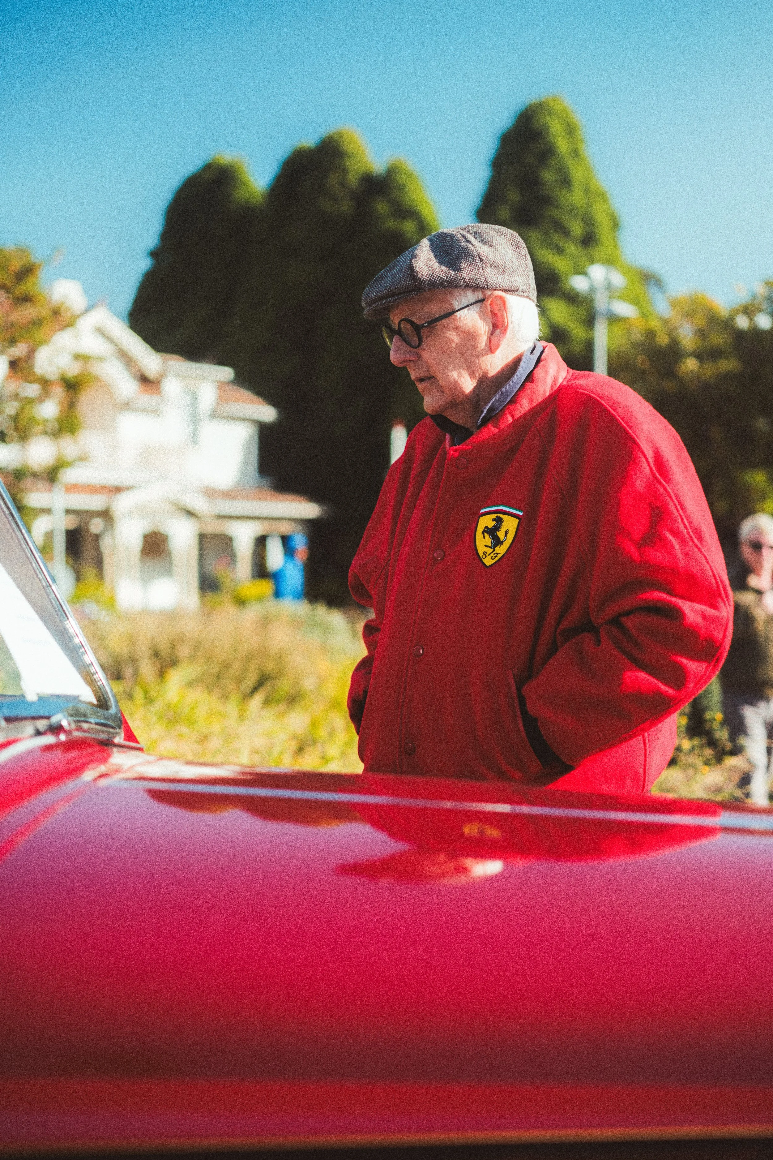 An elderly man with white hair, wearing black-rimmed glasses, a flat cap, and a red jacket with a Ferrari emblem, is looking at a red classic car during daytime.