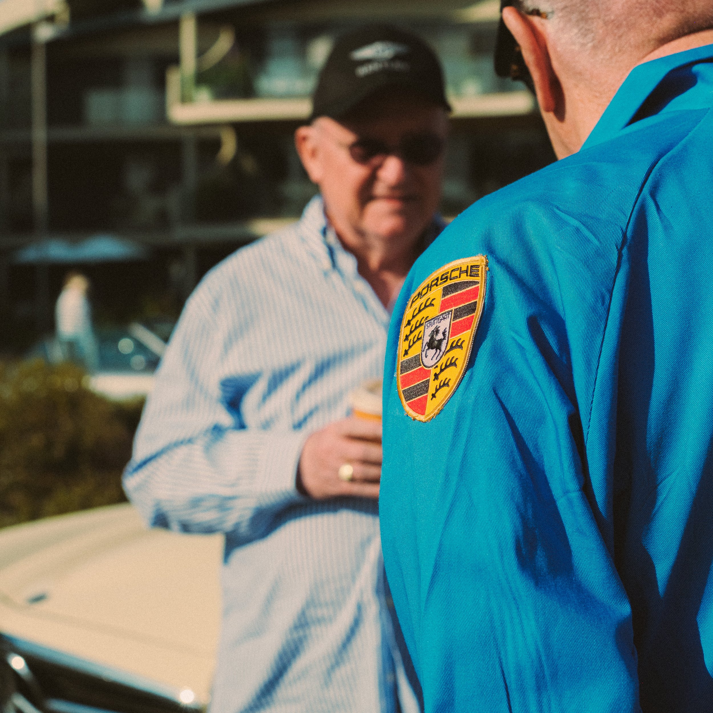 Close-up of a person wearing a blue jacket with a Porsche badge, with another person in the background wearing sunglasses and a striped shirt in a garage or storage area.
