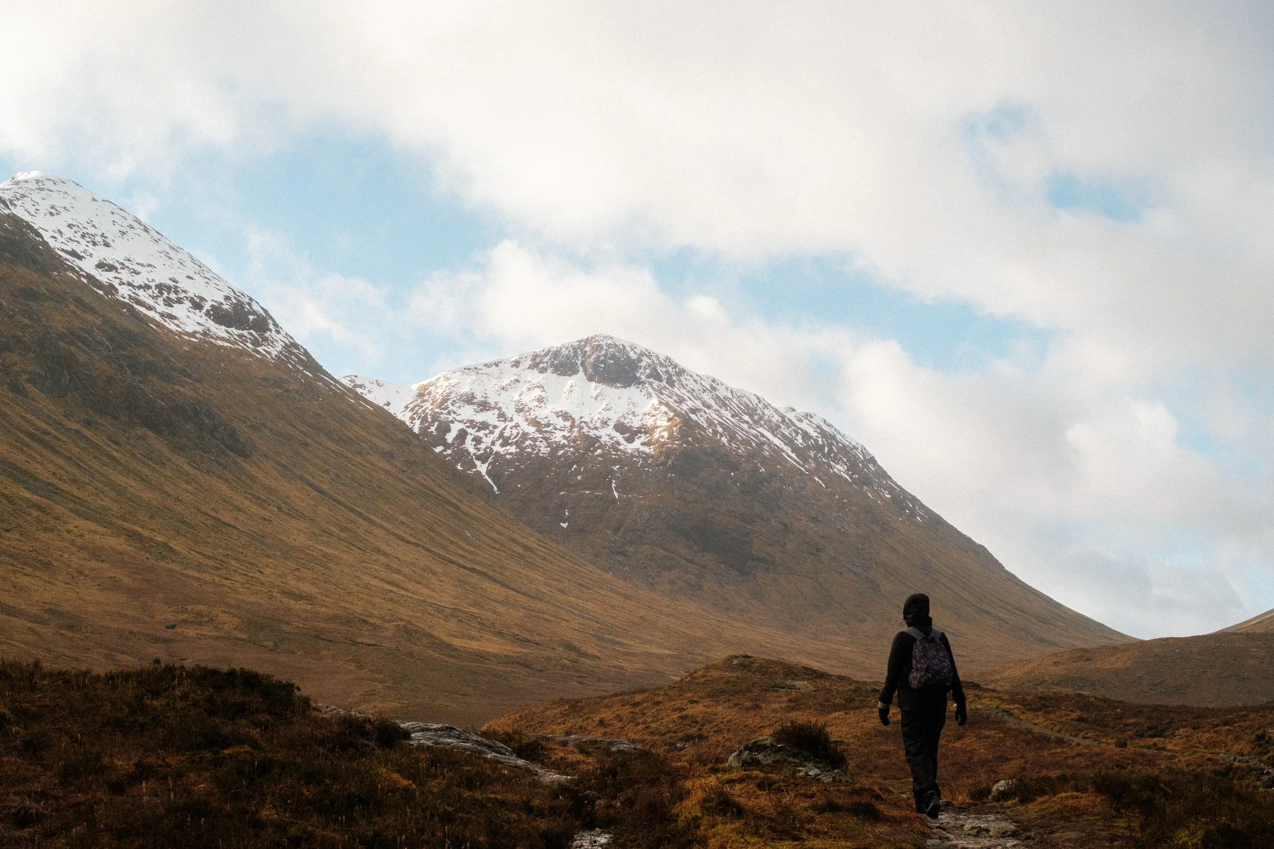 A person hiking through a mountain landscape with snow-capped peaks under a partly cloudy sky.