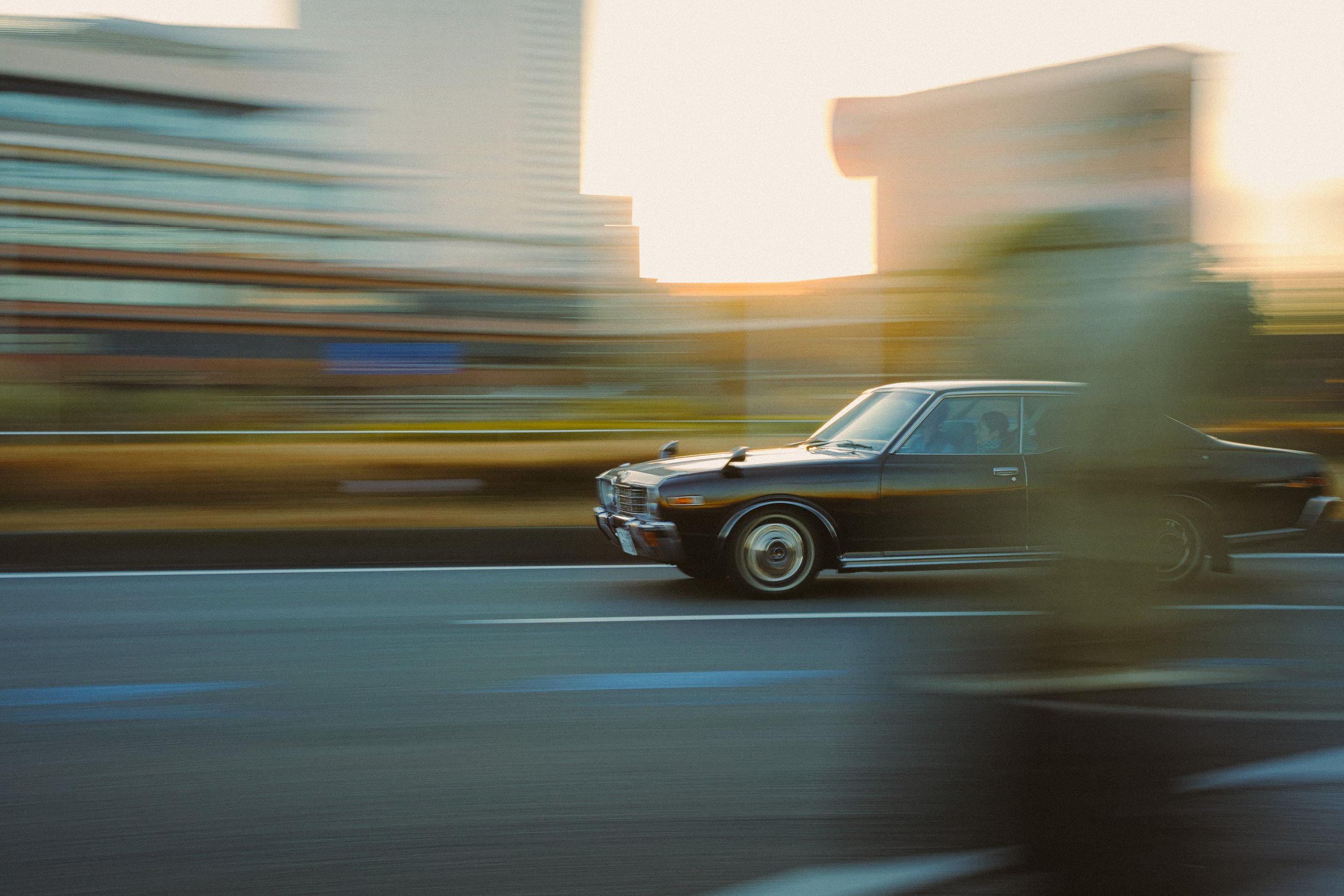A vintage black car driving on a highway with a cityscape of tall buildings in the background during sunset.