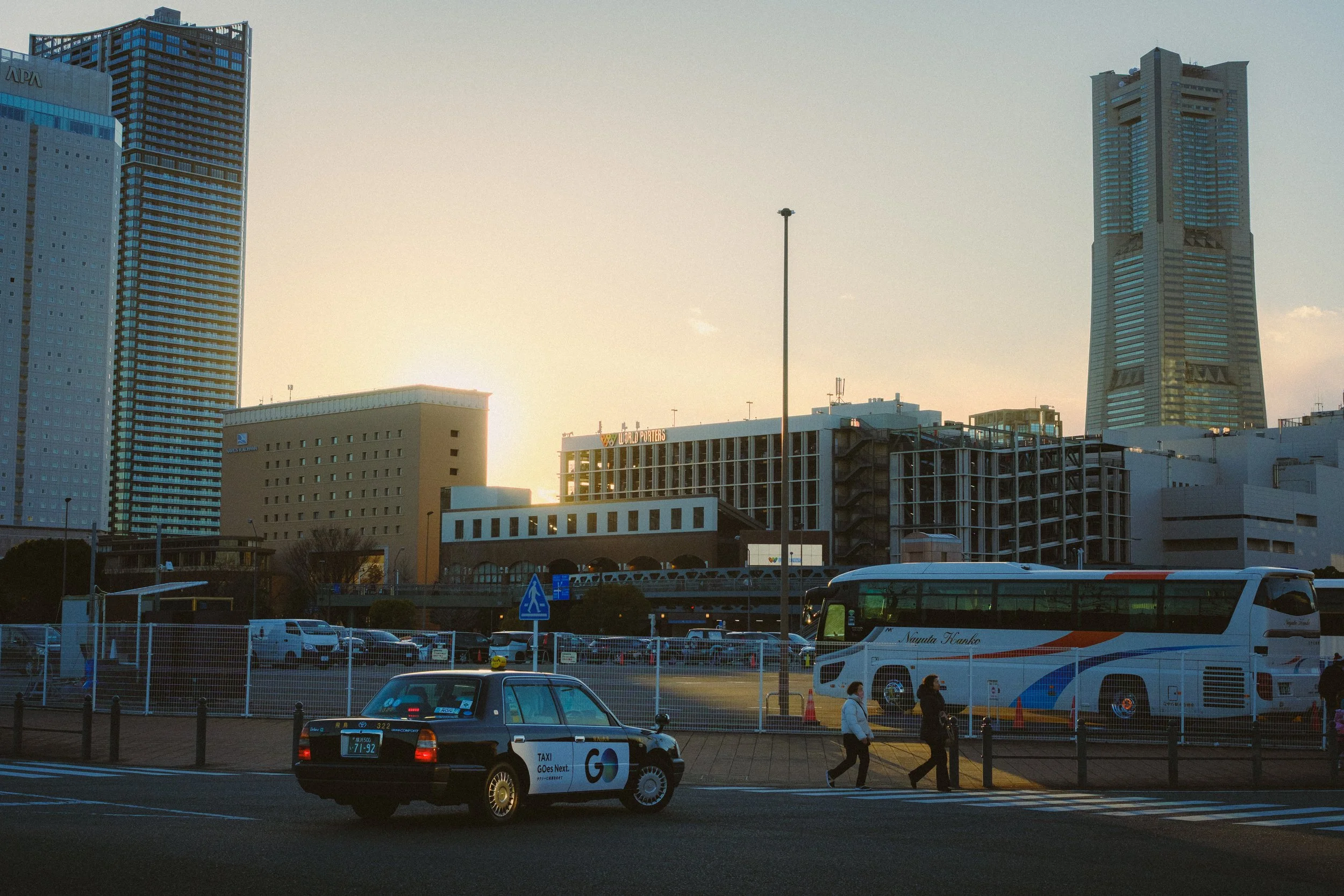 Cityscape with tall buildings during sunset, including a building with the 'APA' logo, a distinctive skyscraper, parked cars, and people walking on the street.