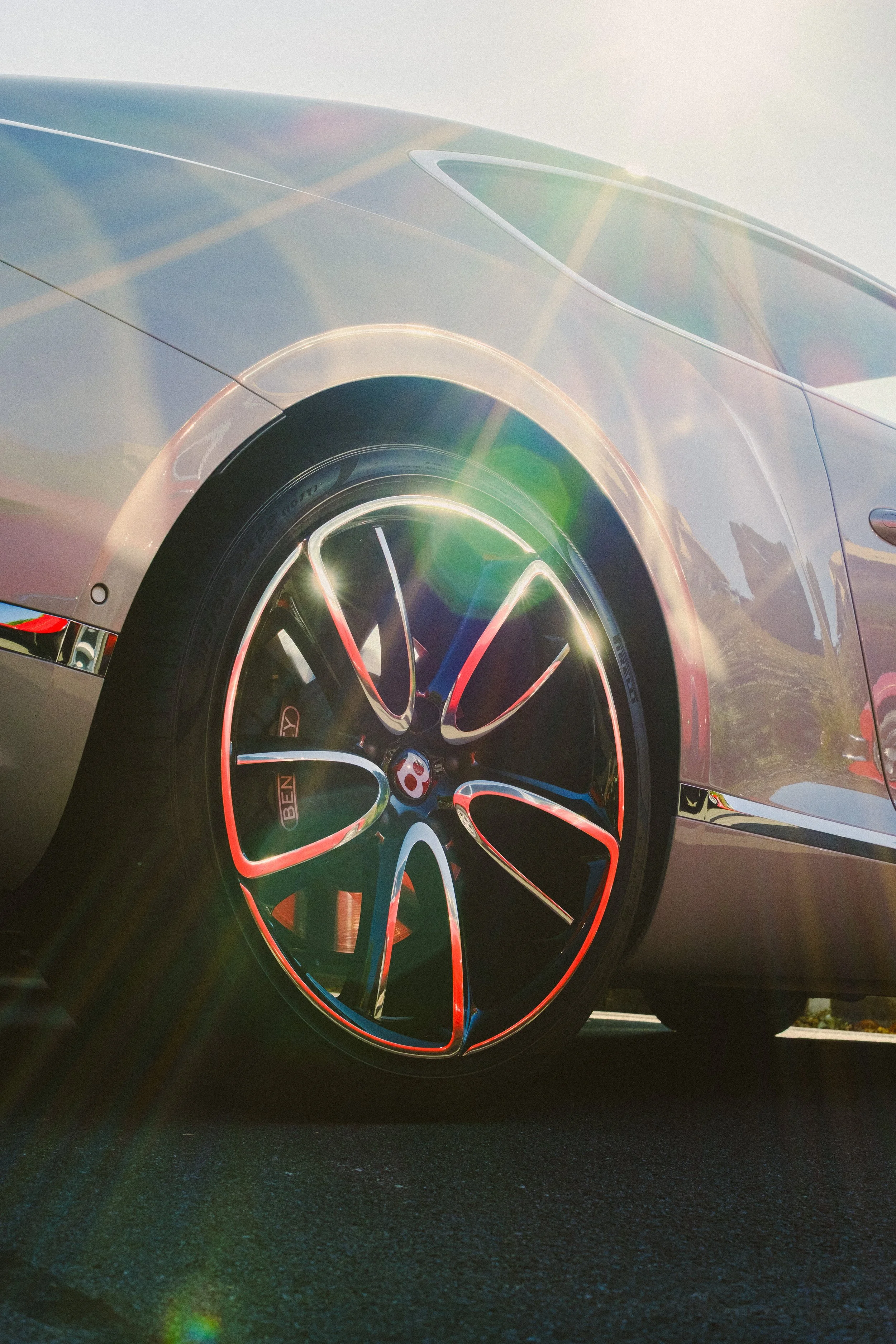 Close-up of a luxury sports car's front wheel and fender, with sunlight glare creating rainbow lens flare effects.
