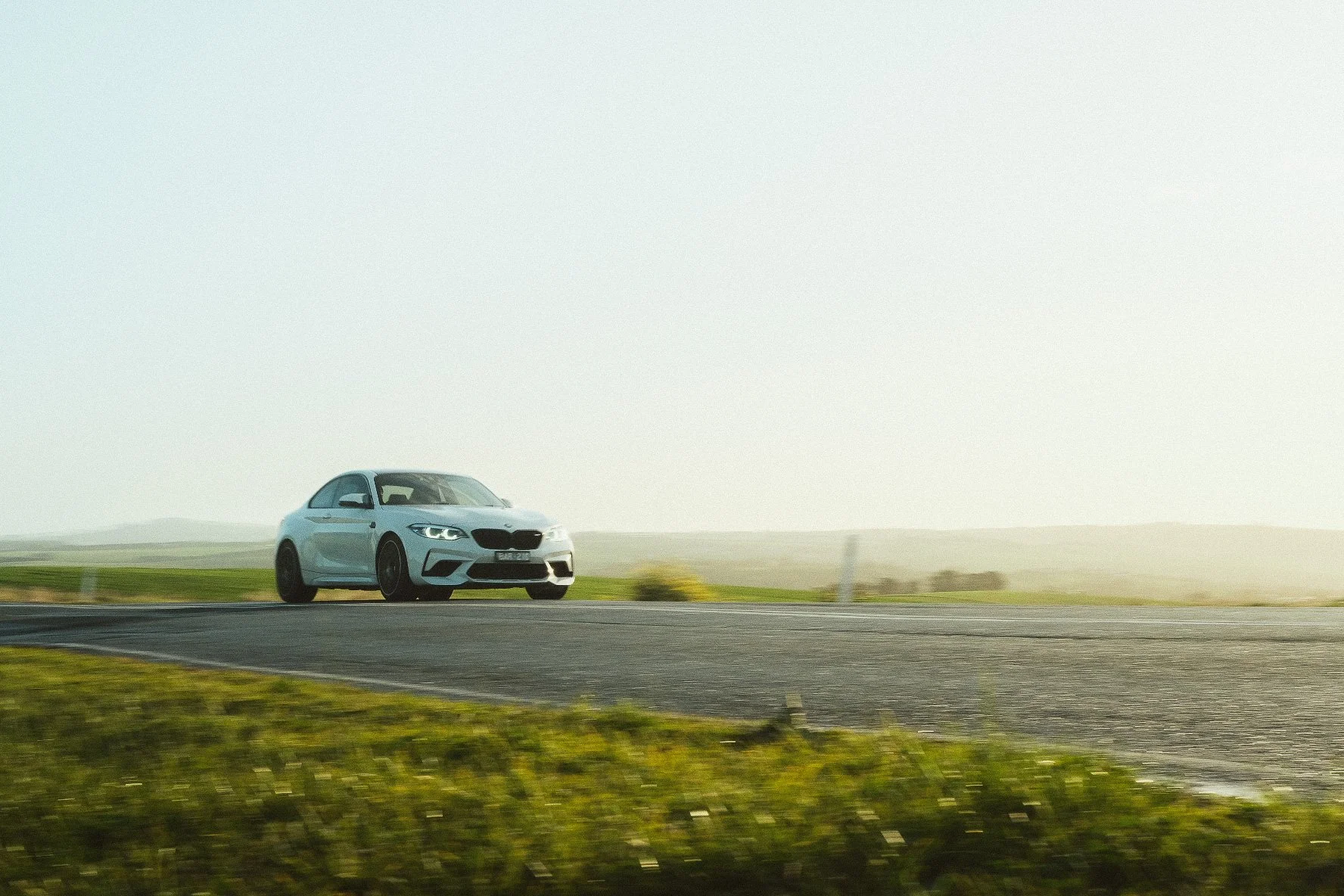 A white car driving on an open road through a grassy landscape with hills in the distance during daylight.