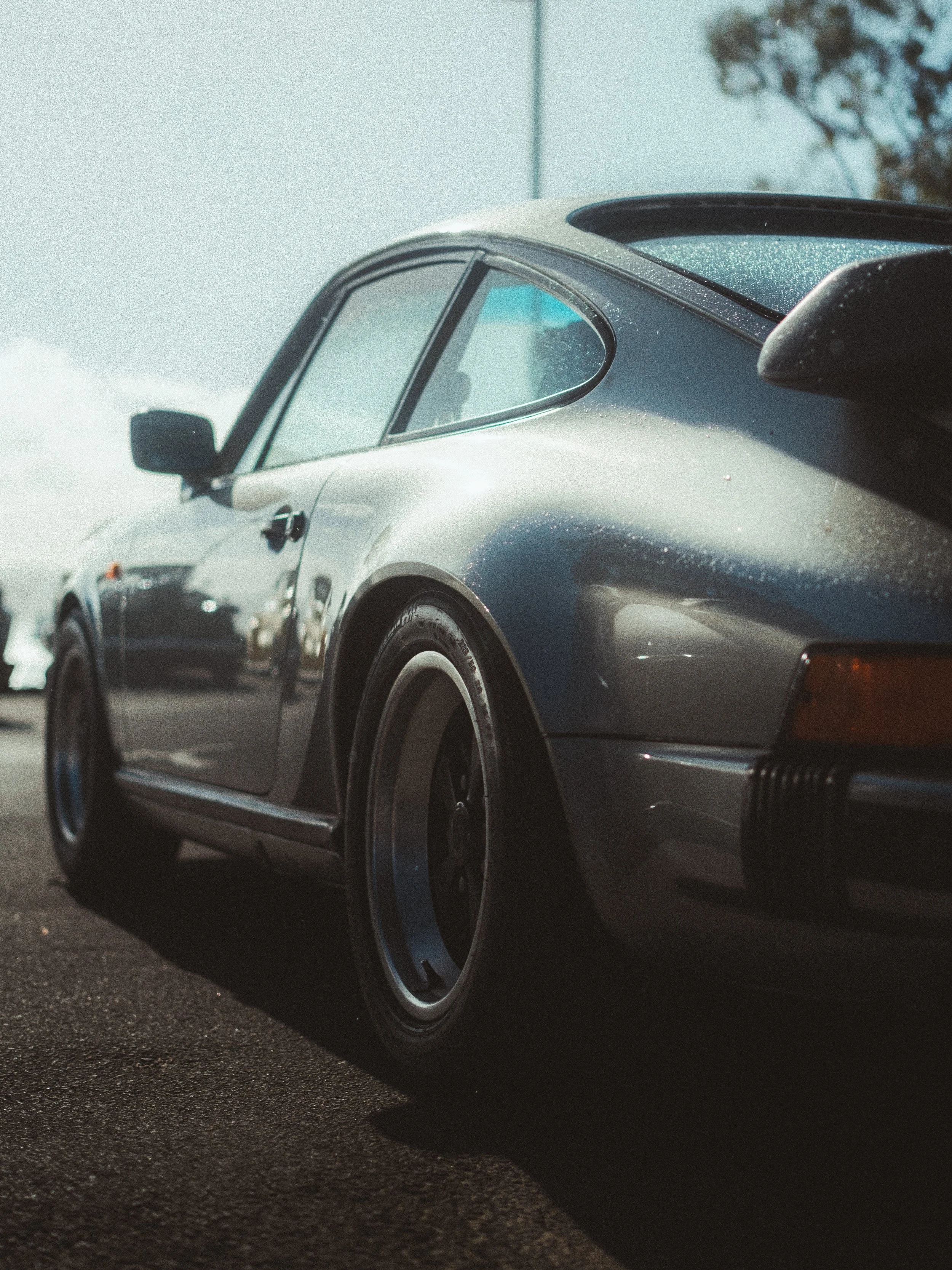 Close-up of a vintage gray Porsche 911 parked on a wet road with raindrops on its roof, side window, and bumper, backlit by a clear sky and blurred background trees.