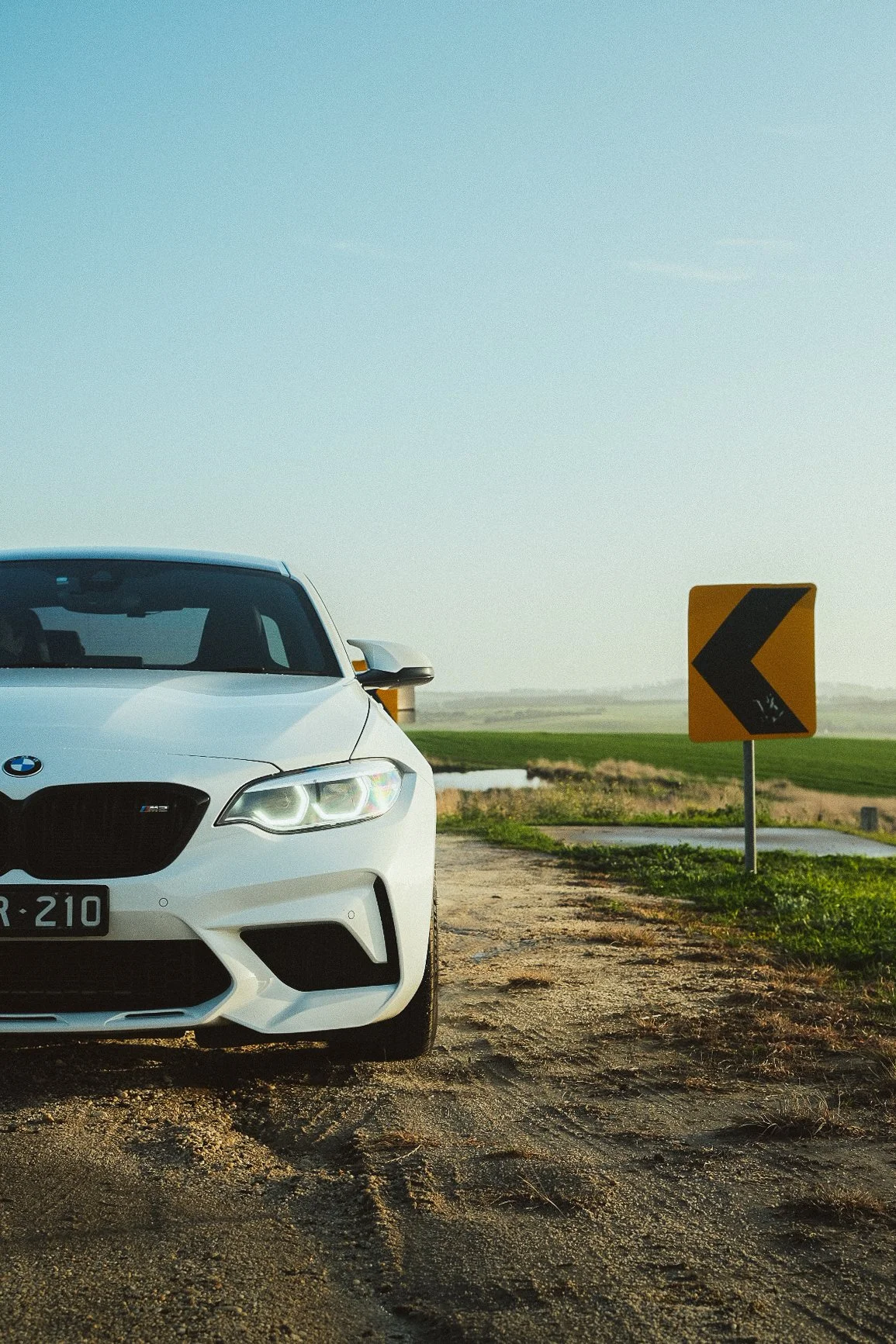 A white BMW M2 parked beside a rural road with a yellow left turn warning sign seen in the background. The scene is set outdoors with flat grassy fields and a partly cloudy sky.