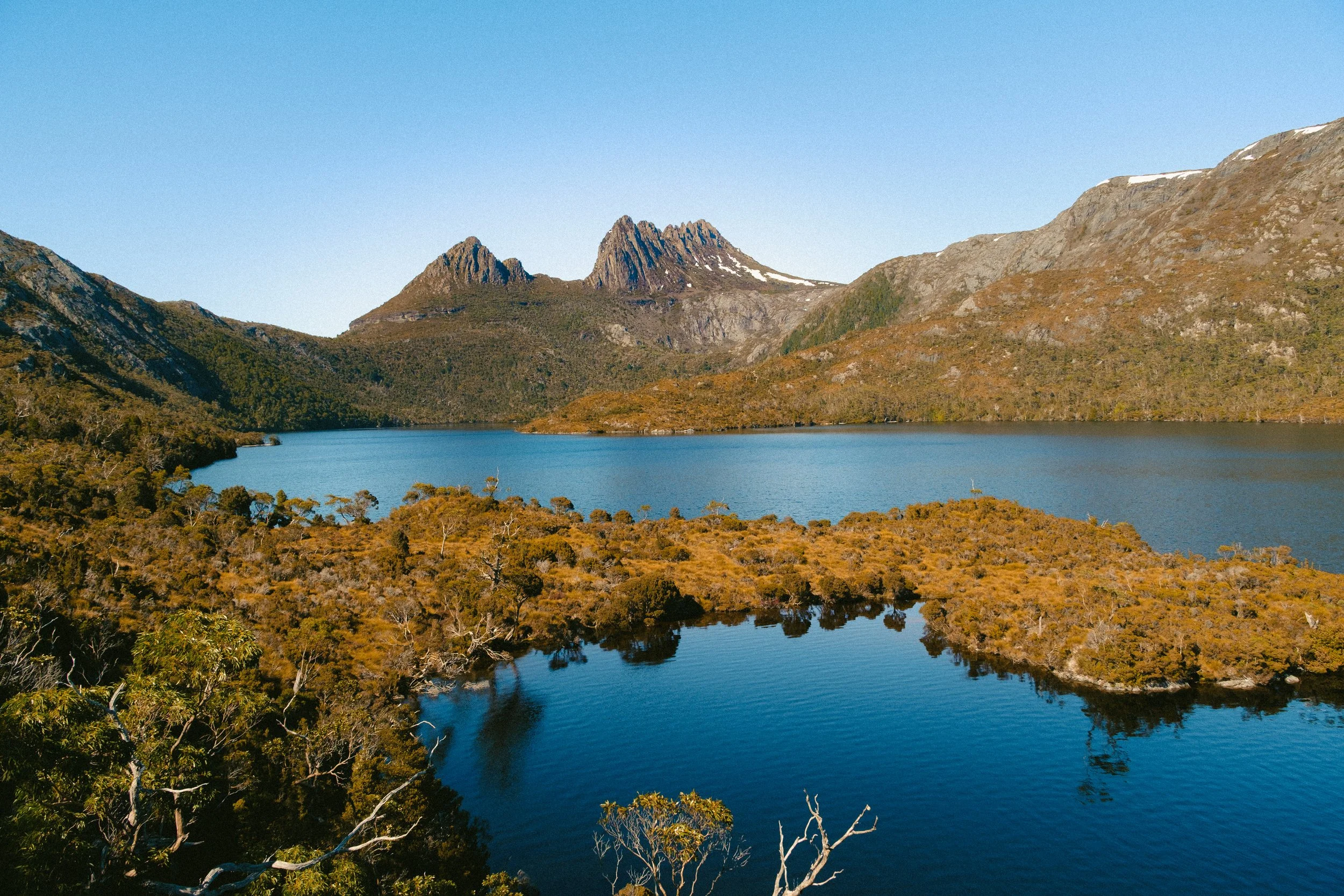 A landscape of a mountain lake with forested shoreline, rugged mountains with patches of snow, and a clear blue sky.