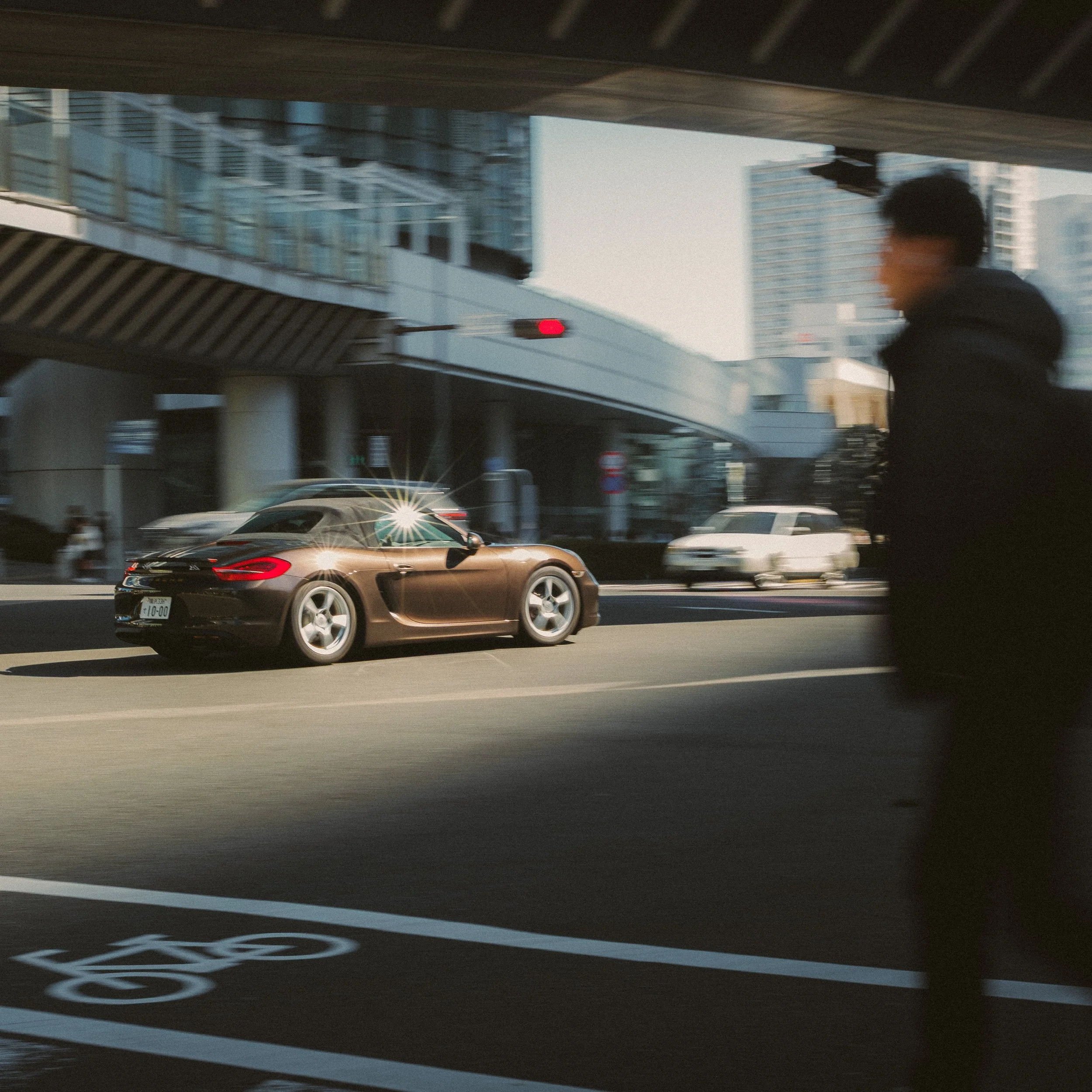 A brown car driving on a city street under an overpass with tall buildings in the background.