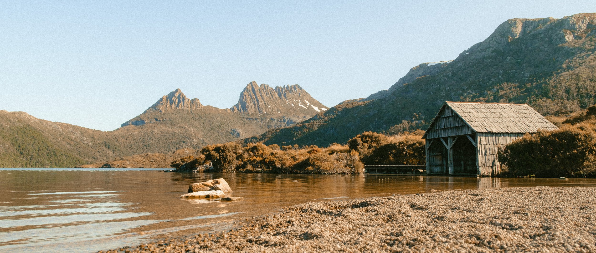 A lakeside scene with a small wooden boathouse, rocky shoreline, calm water, surrounded by mountains and clear sky.