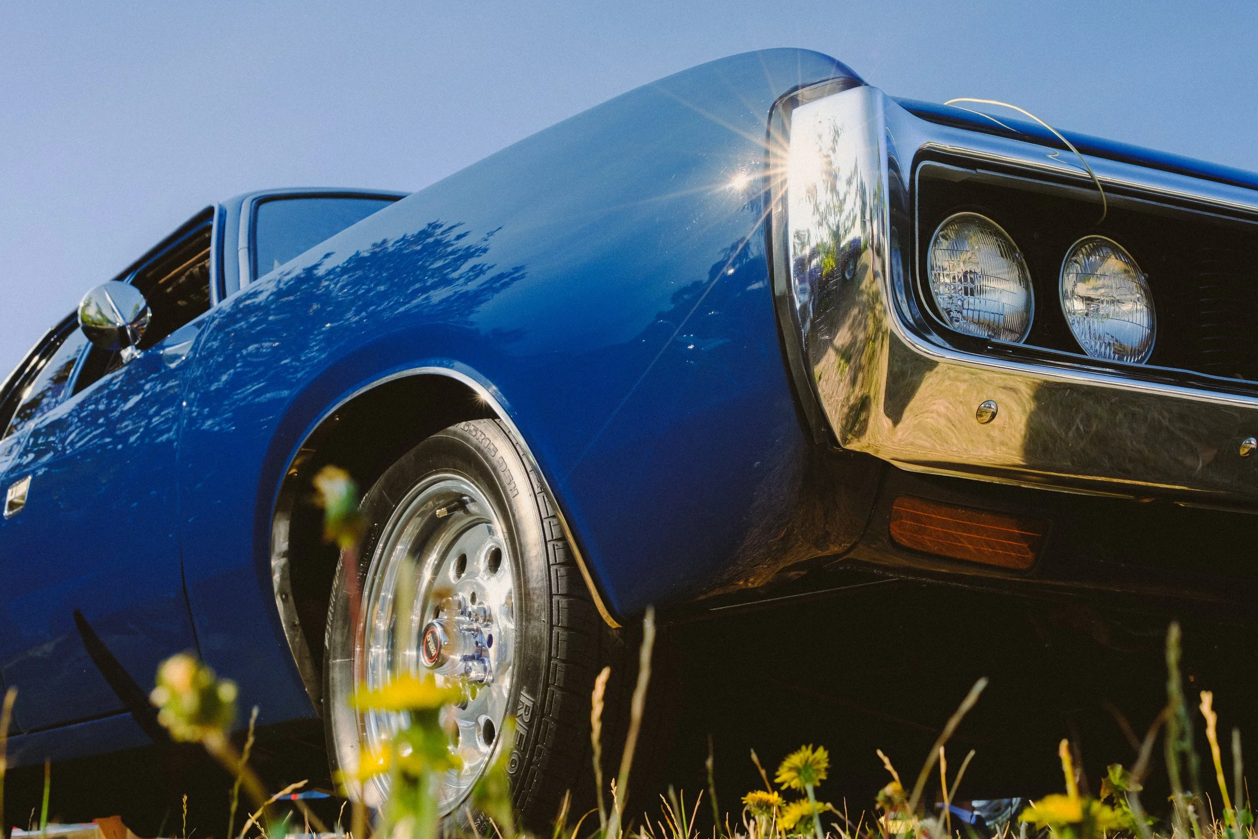 Close-up of a shiny black vintage car with round headlights, chrome bumper, and silver wheel rims, parked on the grass during daytime with blue sky and trees reflected on the car's surface.