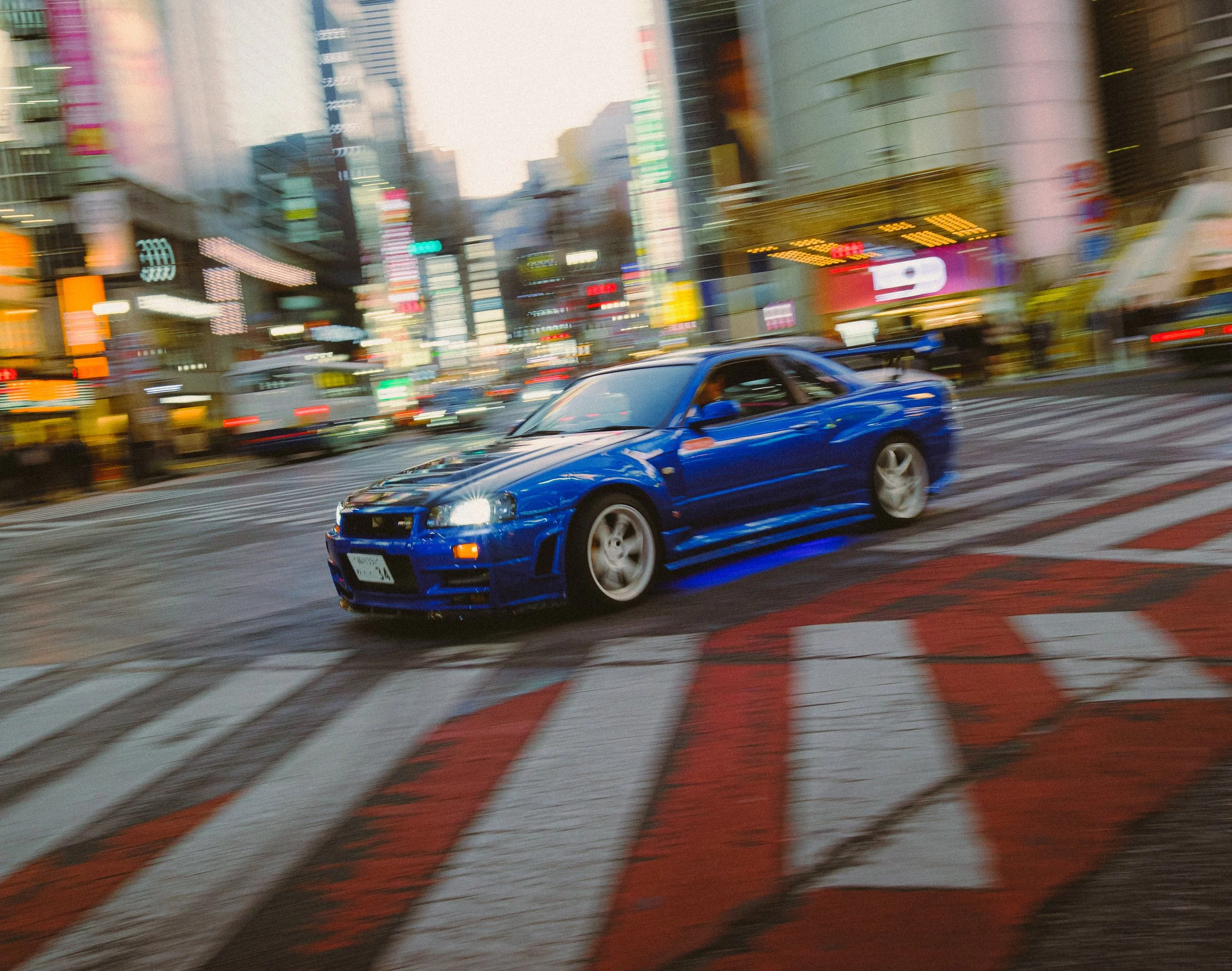 A blue sports car is driving across a city street at night, with bright neon lights and tall commercial buildings in the background. The photo is blurry, suggesting motion.
