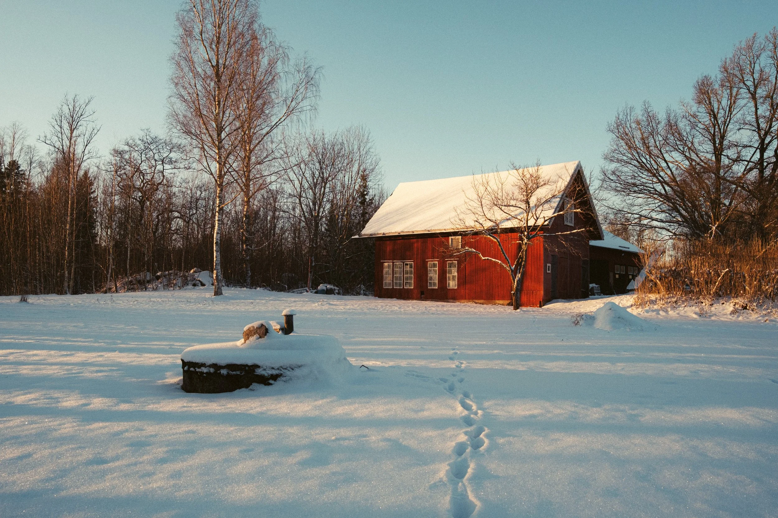 A snow-covered landscape with a red wooden house surrounded by leafless trees under a clear blue sky.