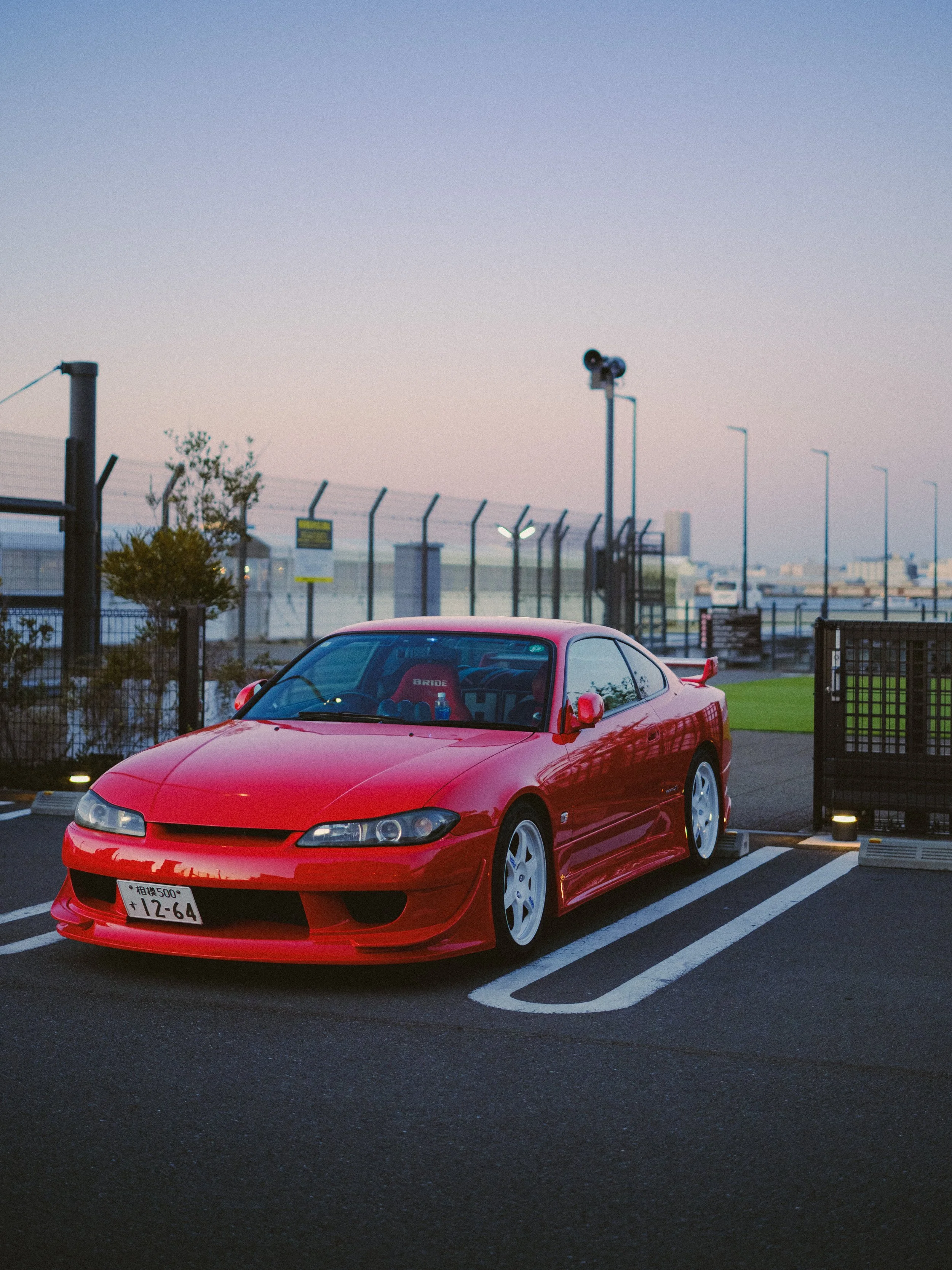 Red sports car parked in a parking lot during dusk, with a fence, security cameras, trees, streetlights, and city buildings in the background.