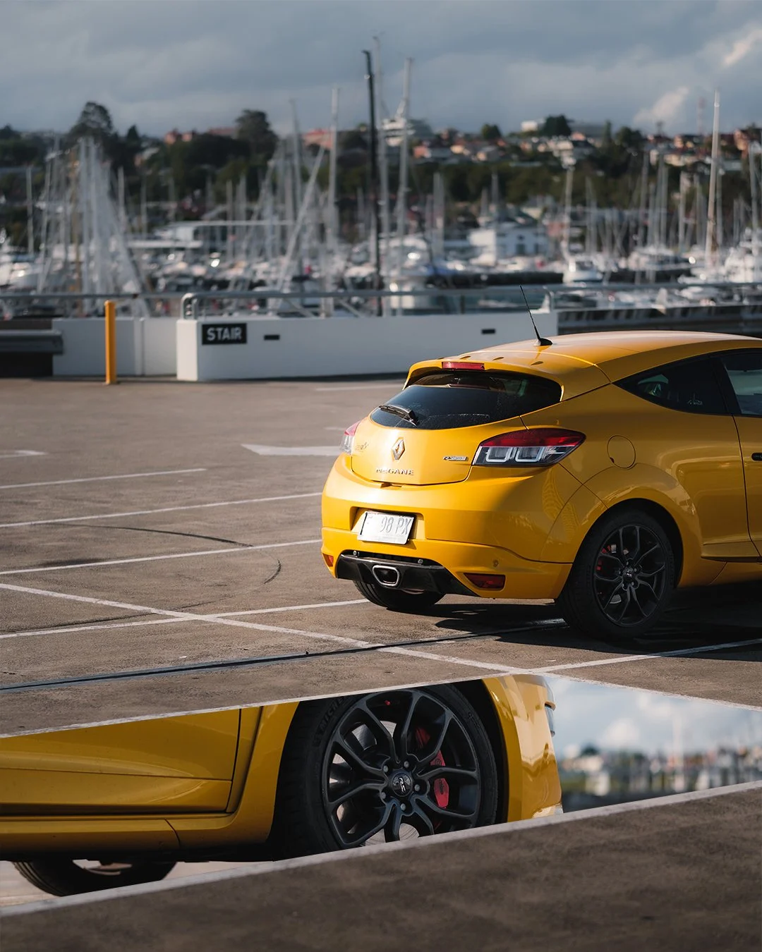 A yellow Renault Megane parked in a parking lot with a marina and sailboats in the background.
