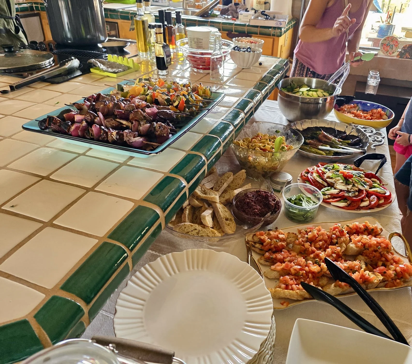 Buffet spread on a tiled kitchen counter with grilled vegetable and meat skewers, caprese salad, bruschetta, pasta salad, and fresh bread.