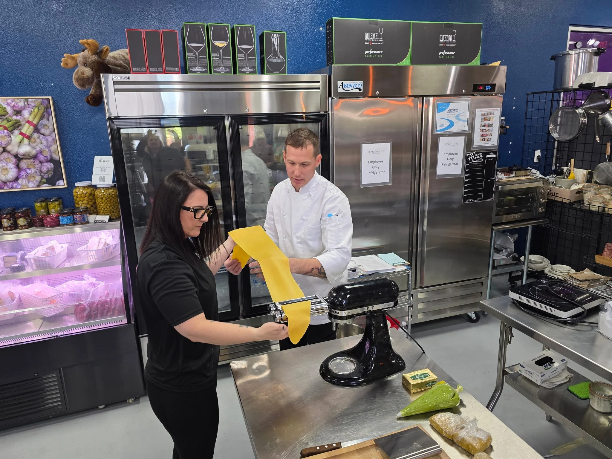 Chef Mitch Bell guiding a guest through rolling fresh pasta dough on a stand mixer attachment during a cooking class.
