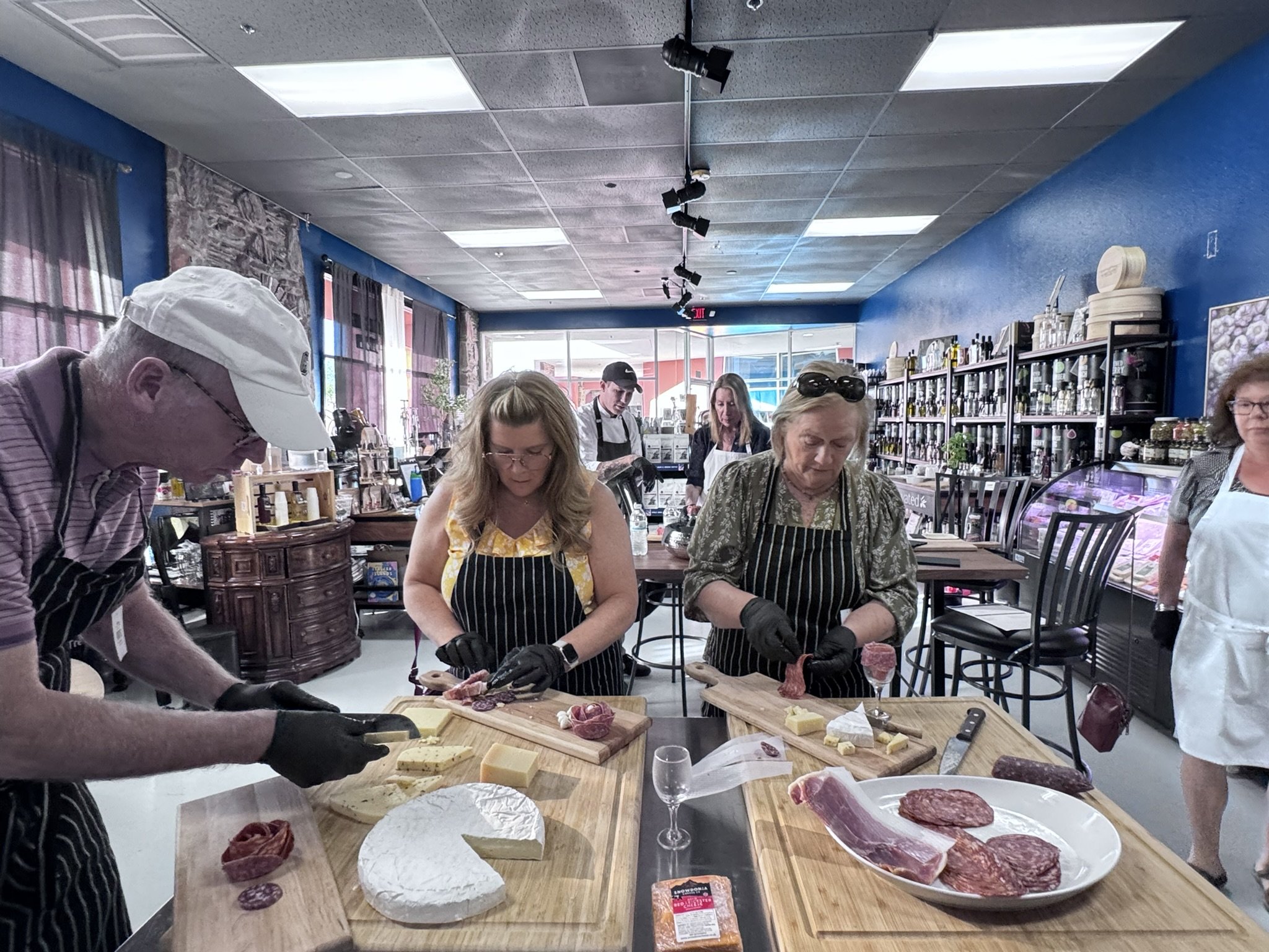 Guests slicing cheese and charcuterie at a hands-on board-building workshop hosted in a specialty foods shop.