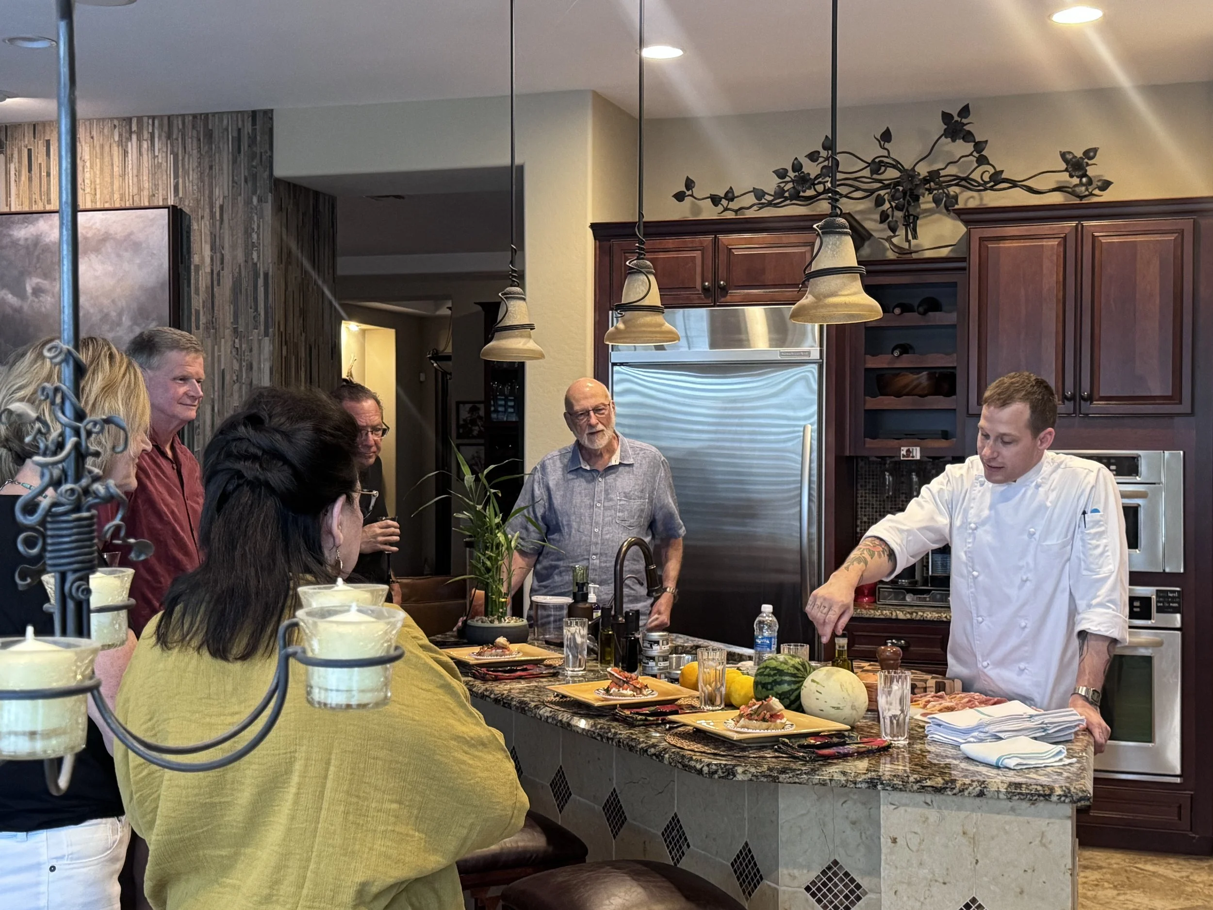 Chef Mitch Bell presenting plated appetizers to a group of guests gathered around a kitchen island during an in-home dining event.