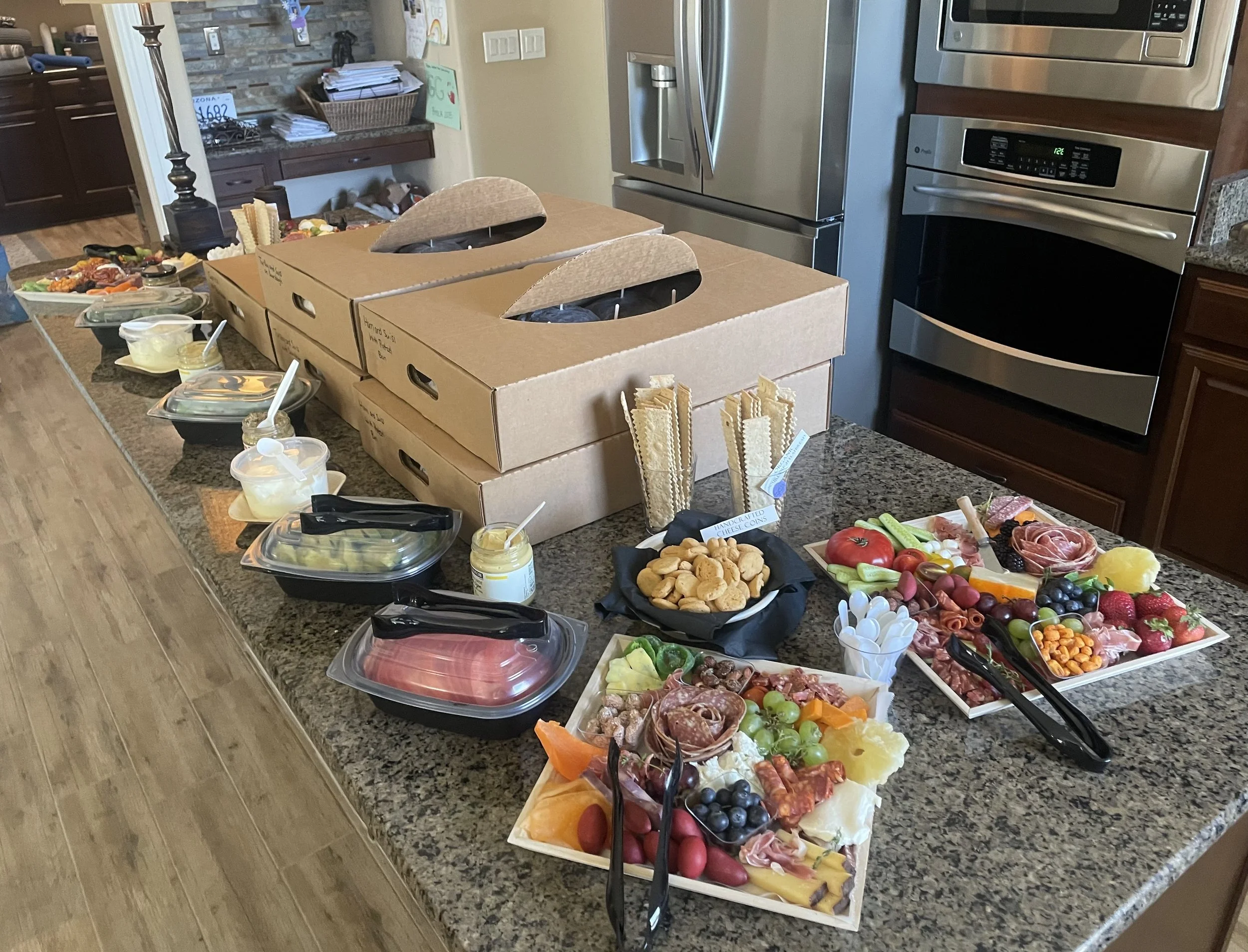 Kitchen island catering spread with multiple charcuterie boards, meat and cheese trays, crackers, and packaged sandwich boxes for a private event.