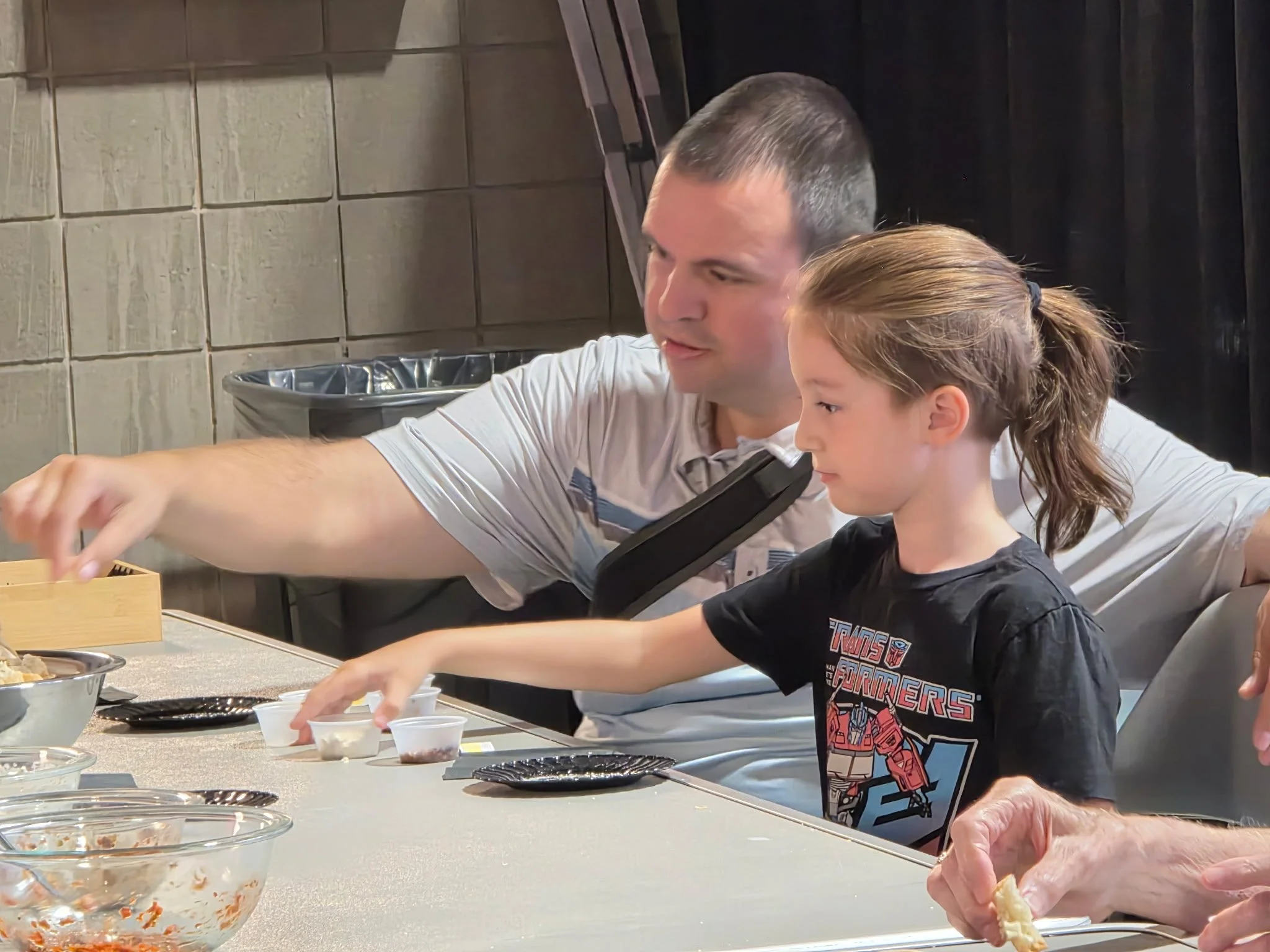 A father and daughter reaching for sample cups at a live cooking demo audience table.