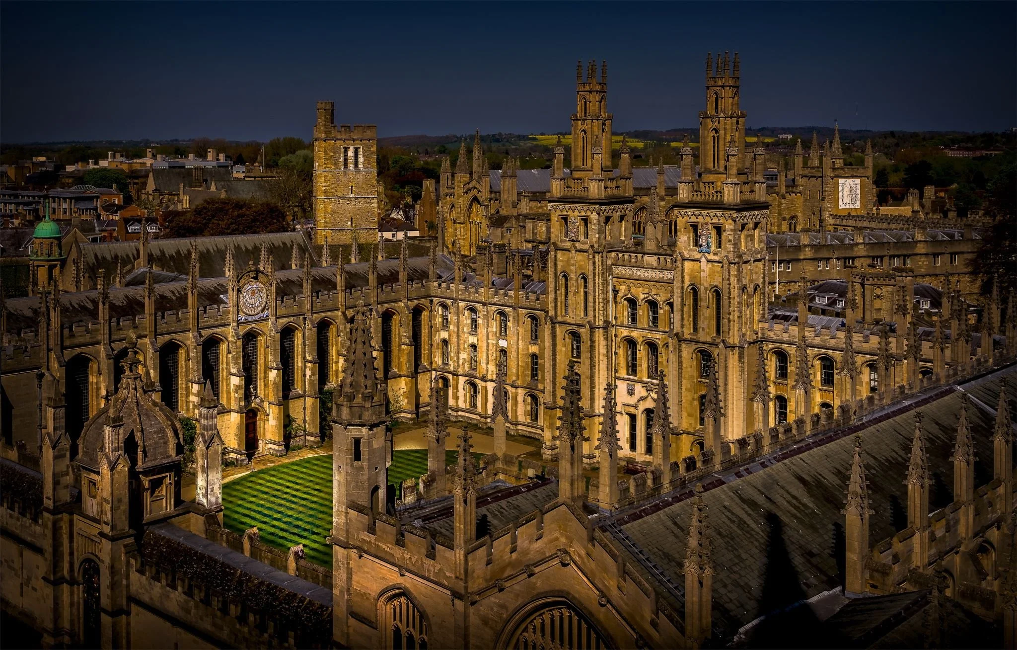 A night time aerial view of All Souls College, showing the chapel, the Hawksmoor towers and the Codrington Library, set against a dark sky with some distant landscape in the background.