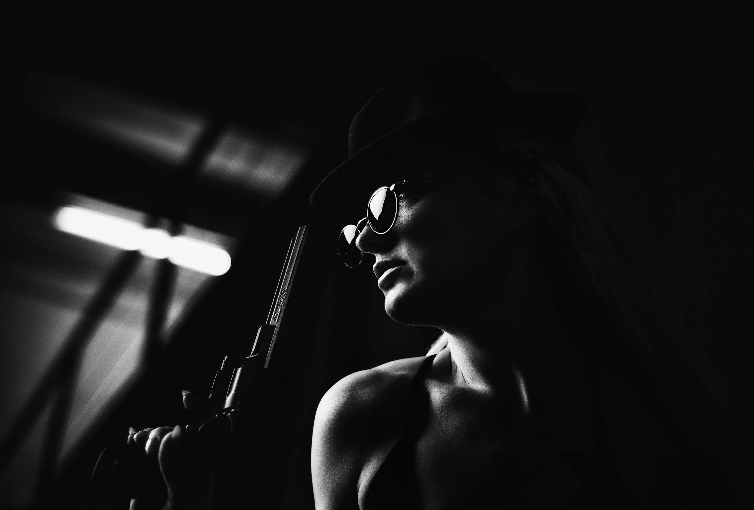 A person wearing sunglasses and a hat, holding a handgun in a dark setting.