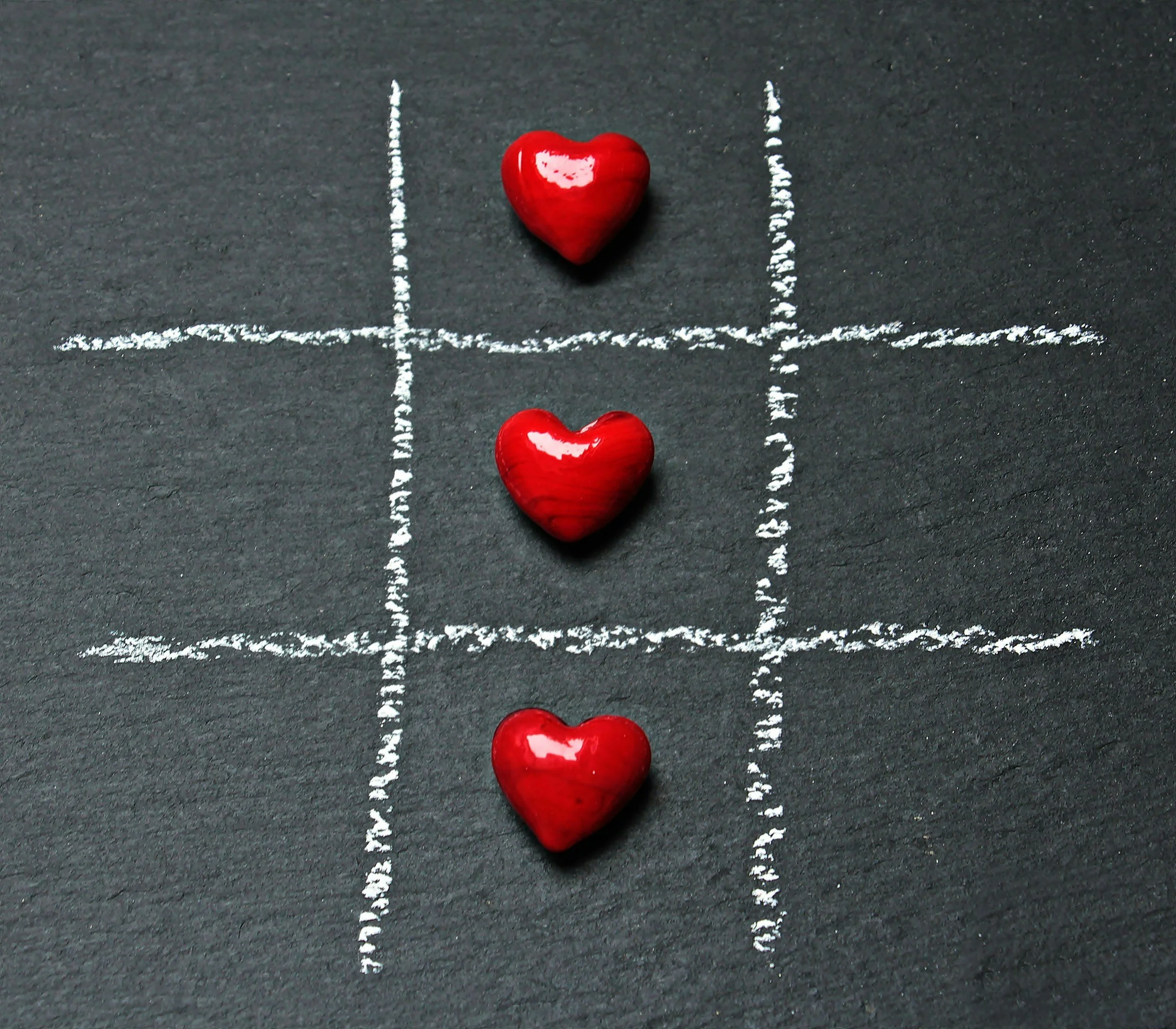 Three red hearts and white chalk lines on a black surface, forming a tic-tac-toe game with hearts as the 'X's.