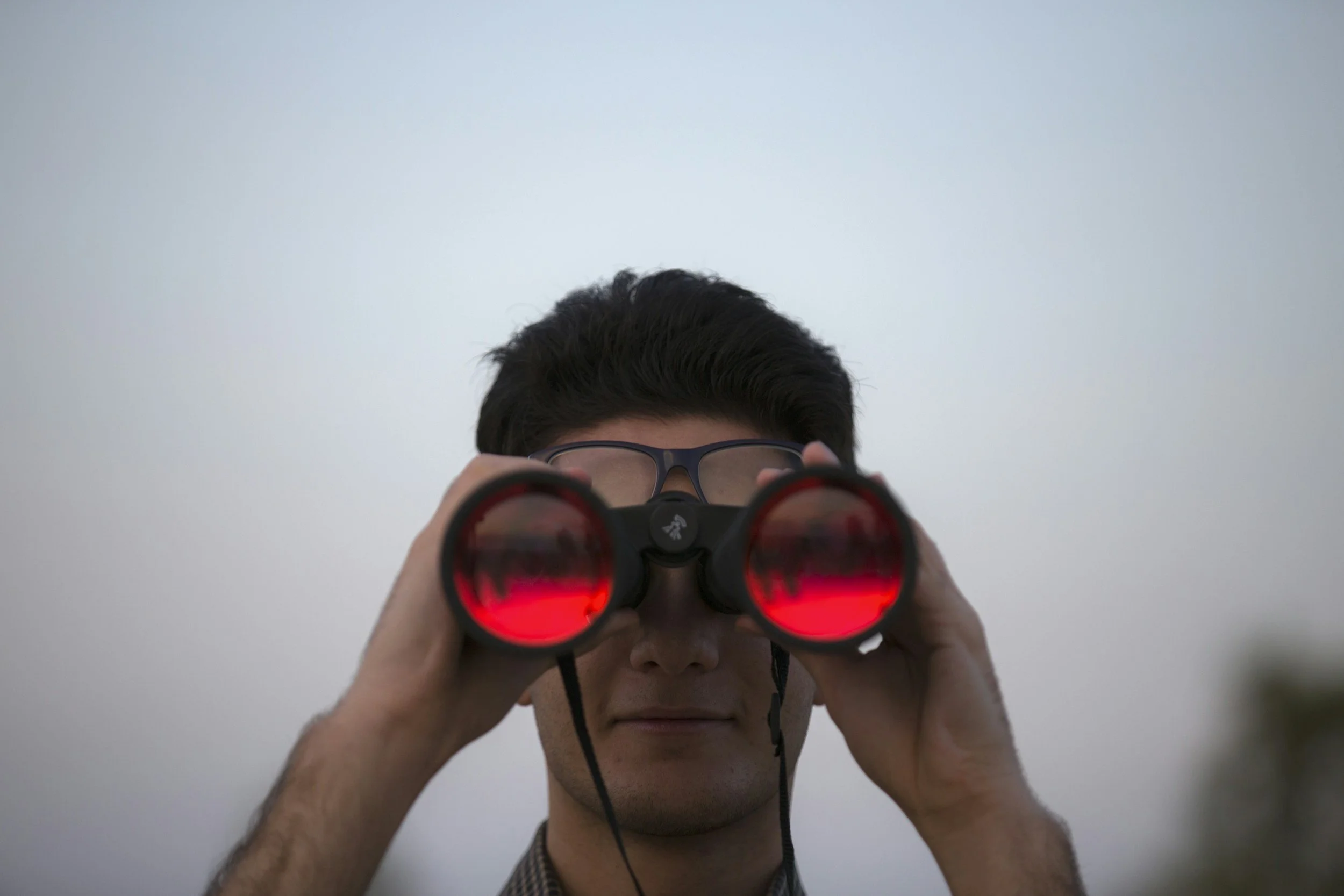 Person looking through red binoculars, partial view of face, wearing glasses, background is plain and light-colored.