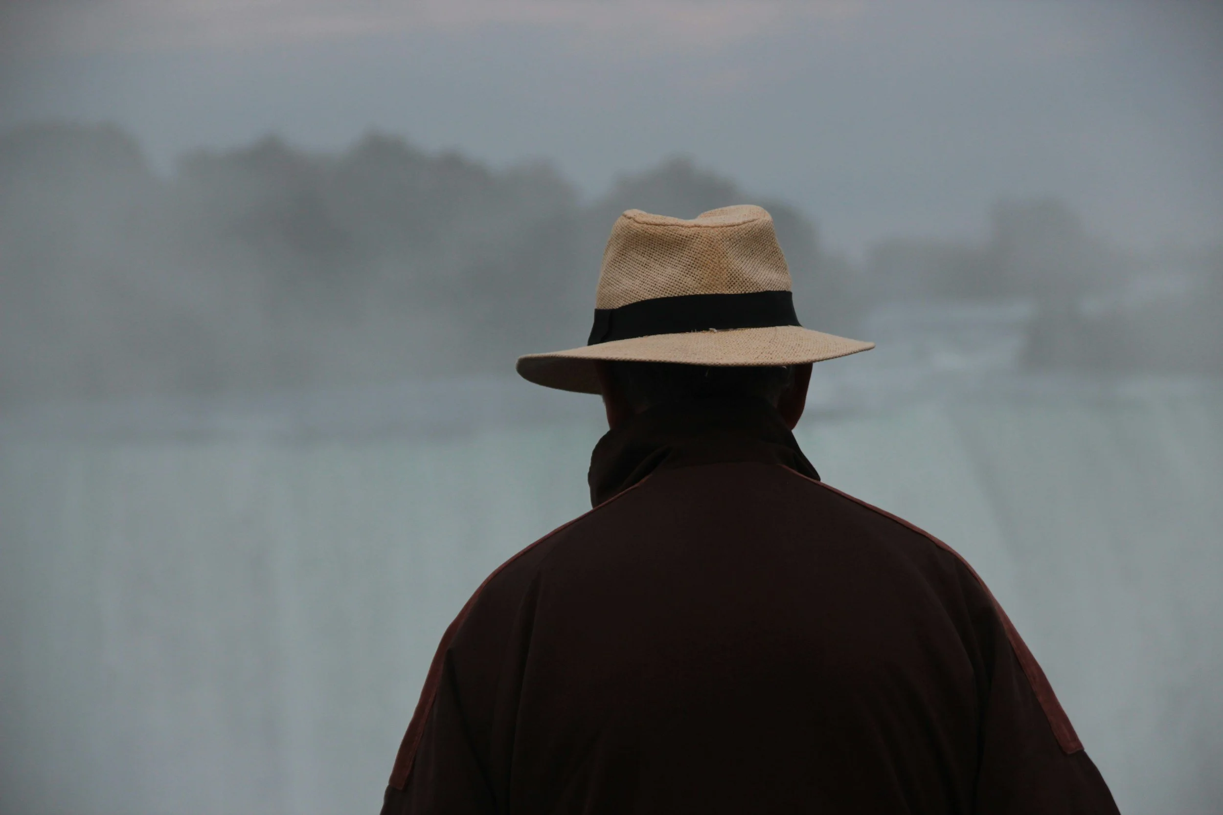 A person wearing a beige wide-brimmed hat and a dark jacket, facing away and looking out at a gray ocean or lake with a cloudy sky.