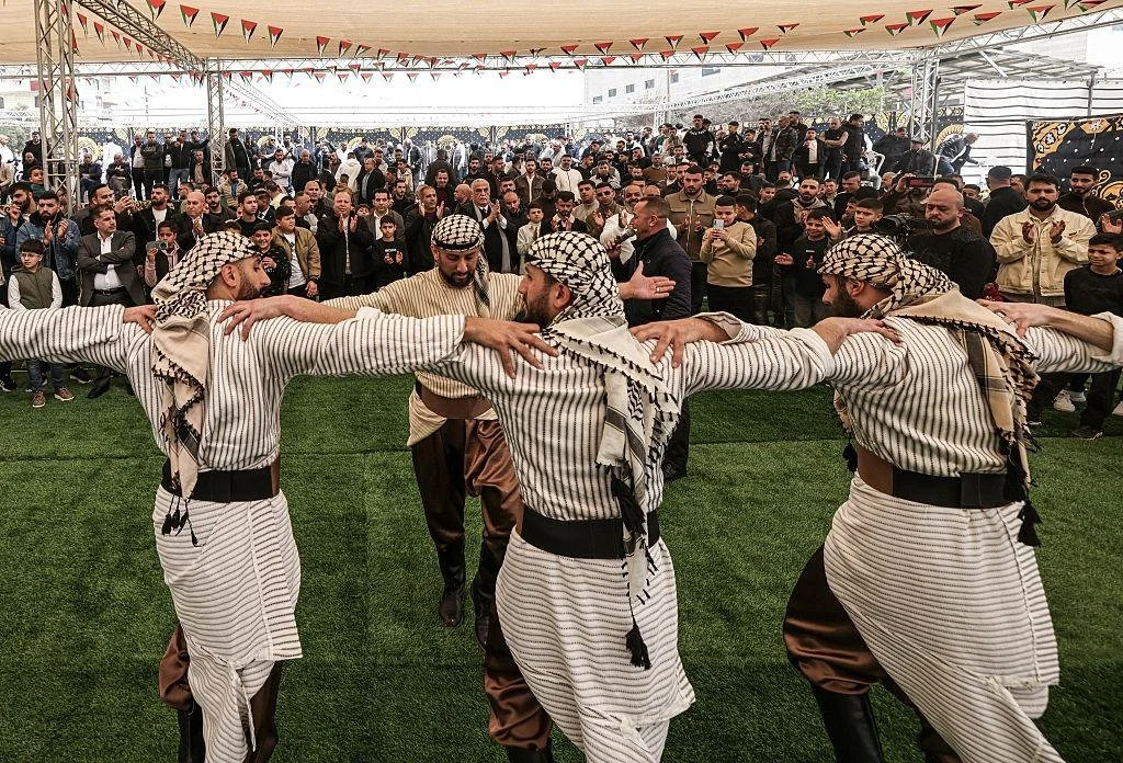 Men in traditional Middle Eastern attire performing a folk dance in a tented outdoor setting with a large audience watching.