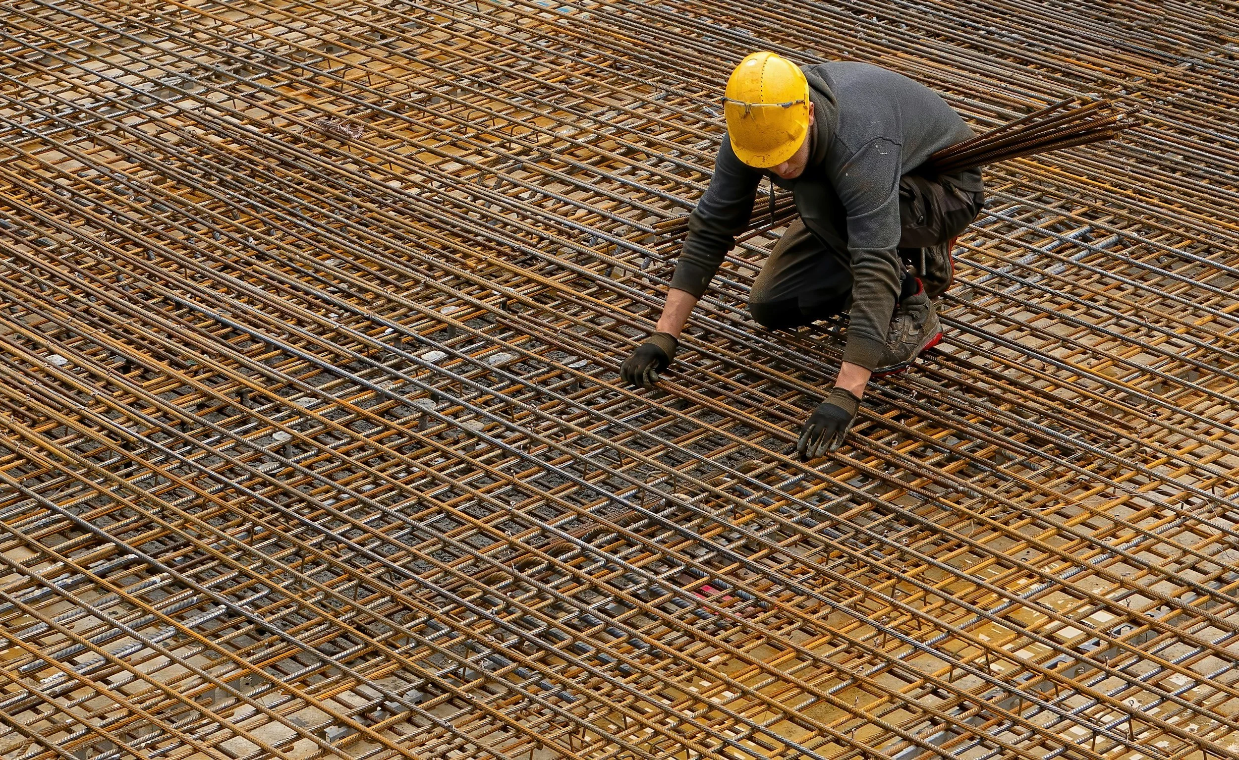 A construction worker kneeling on a steel rebar grid on a building site, wearing a yellow safety helmet, gloves, and dark clothing.