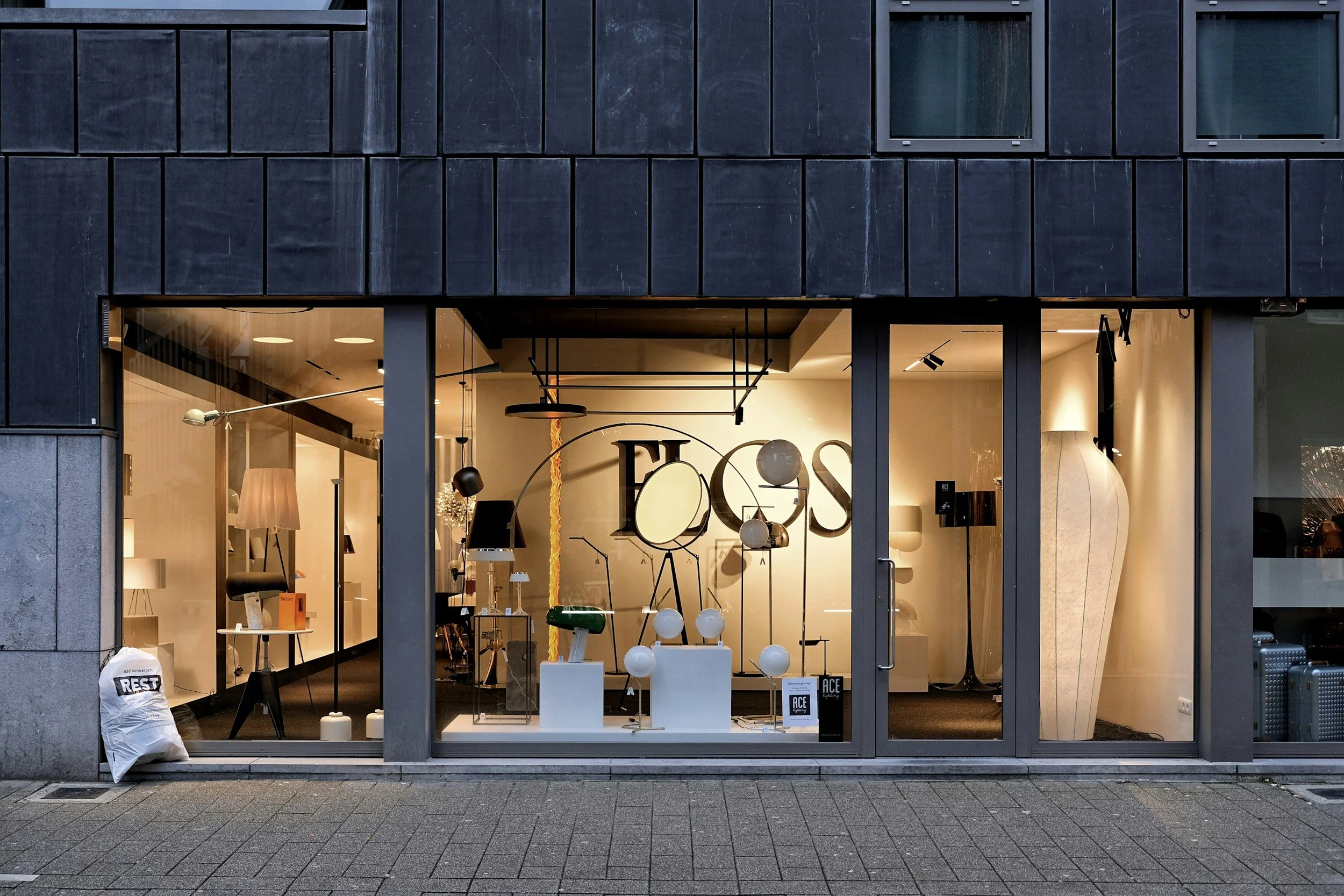 Storefront window display with furniture and lighting fixtures inside, seen through large glass windows and door. Outside, on the sidewalk, there is a white bag labeled 'REST' and a black and white bag.