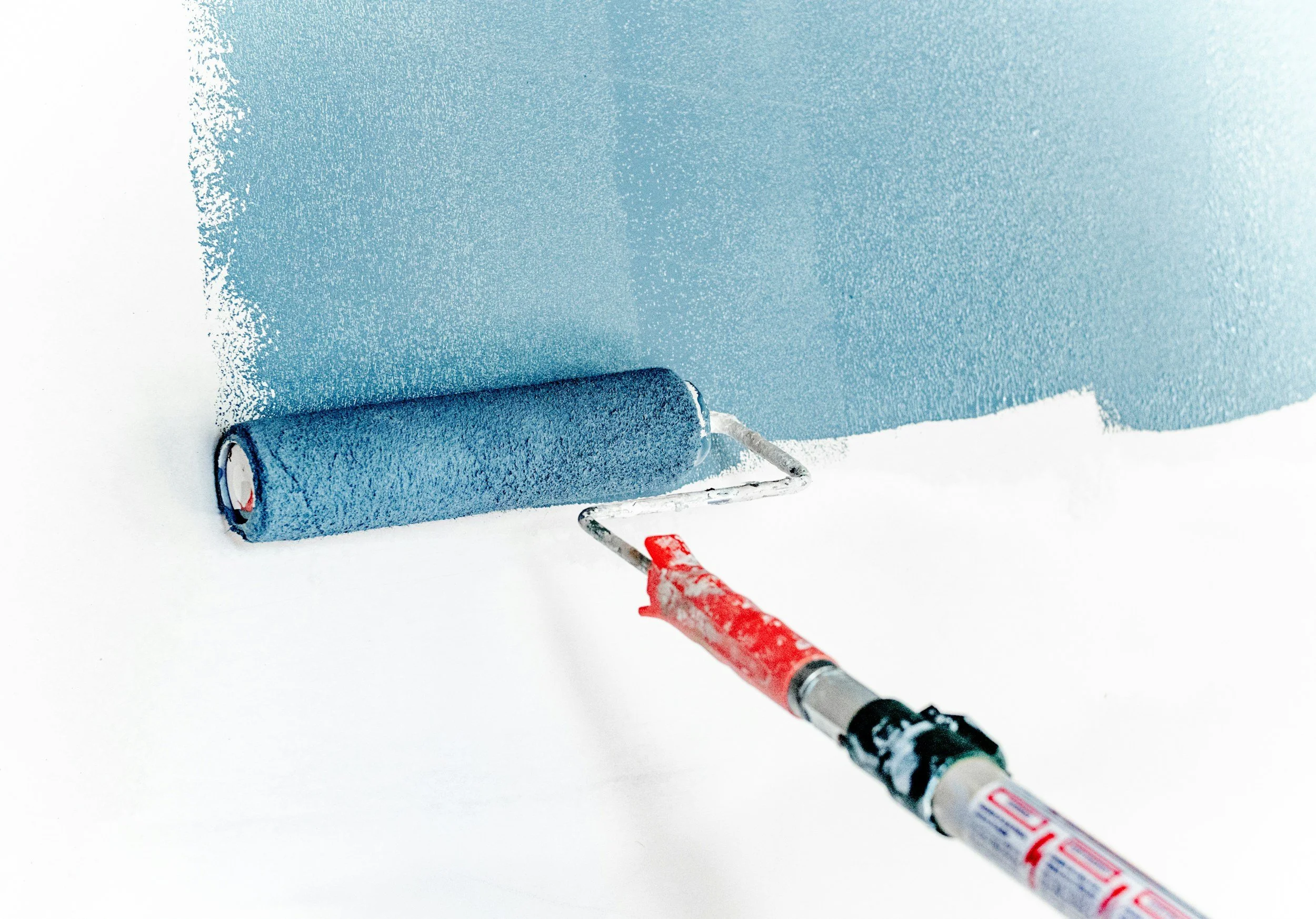 A paint roller covered in blue paint, with a red and white paint sprayer in the foreground, on a white surface.