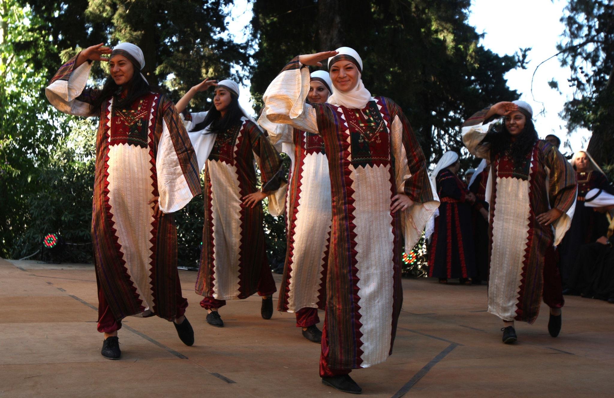 Group of women in traditional dresses performing a dance outdoors, saluting with their right hands to their foreheads, surrounded by trees.