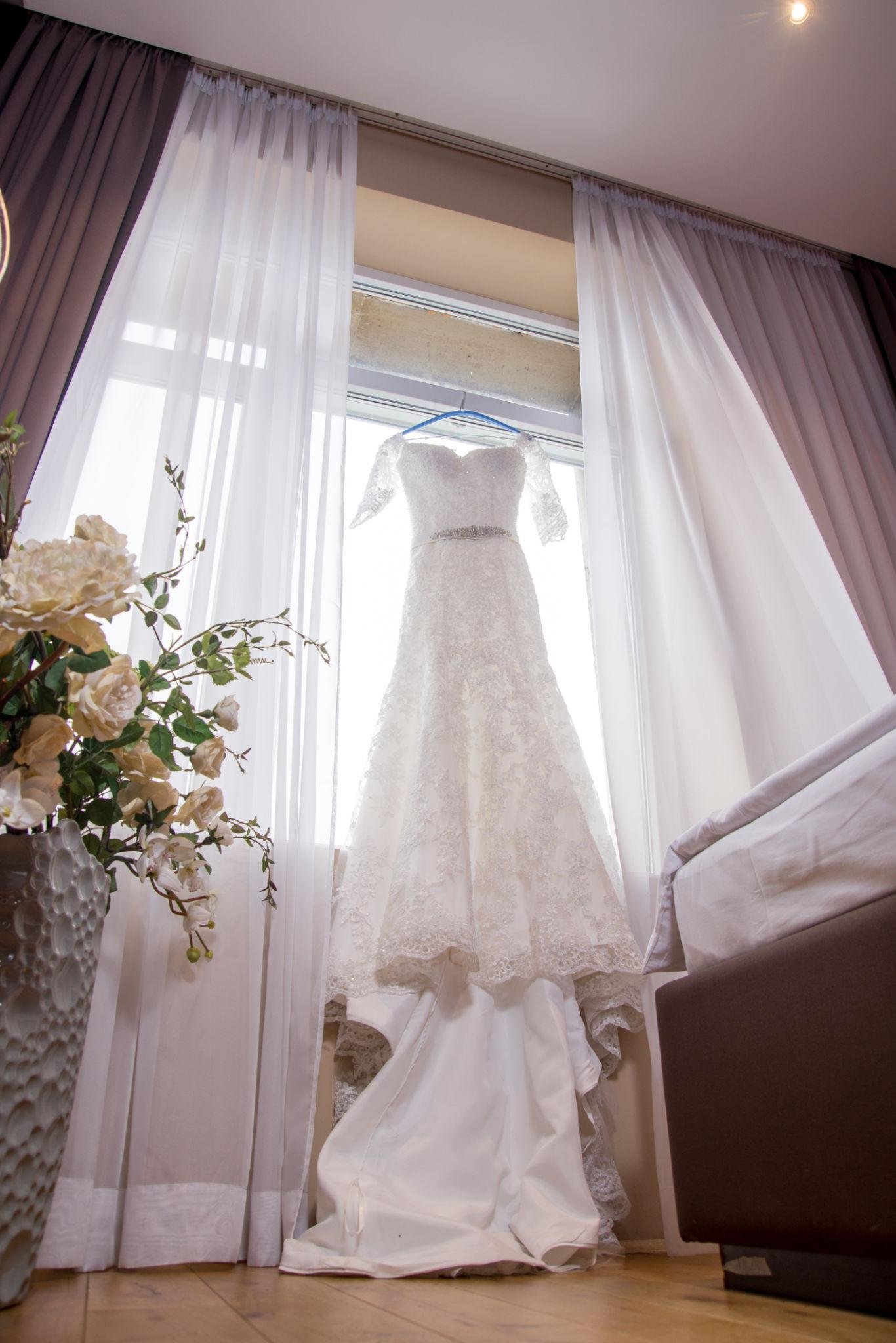 A wedding dress hanging in front of a window with white curtains, next to a vase of white flowers, and a dark armchair nearby.