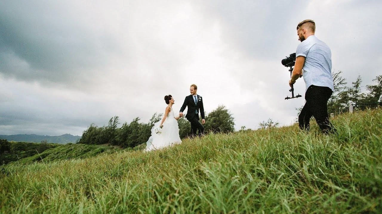 A bride and groom holding hands in a grassy field with cloudy sky, a photographer taking pictures of them.