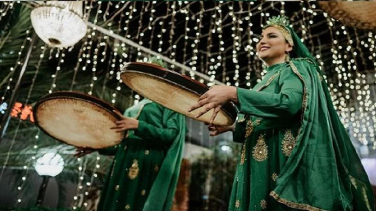A woman dressed in a green traditional outfit, smiling and holding a large round tray during a celebration with festive lights and decorations.