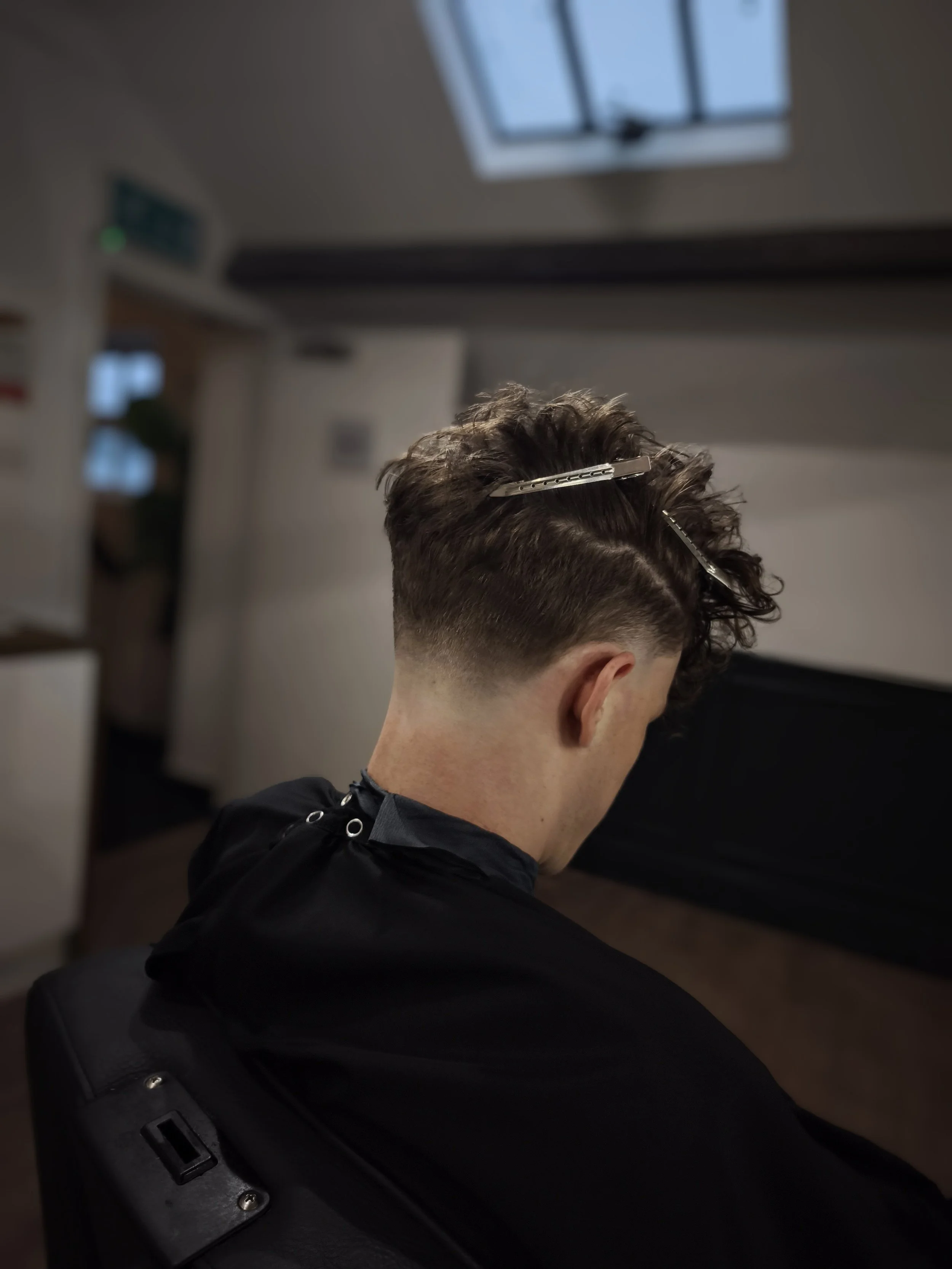 A person with short, curly hair getting a haircut, with hair clips holding sections of hair. The person is seated in a salon chair, wearing a black cape, with a window and ceiling light in the background.