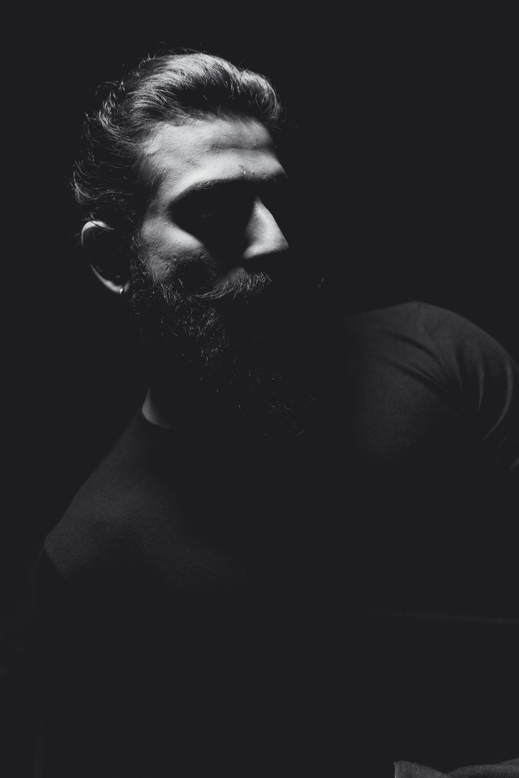 A black and white portrait of a man with a beard, long hair, and a mustache, with lighting highlighting his facial features against a dark background.