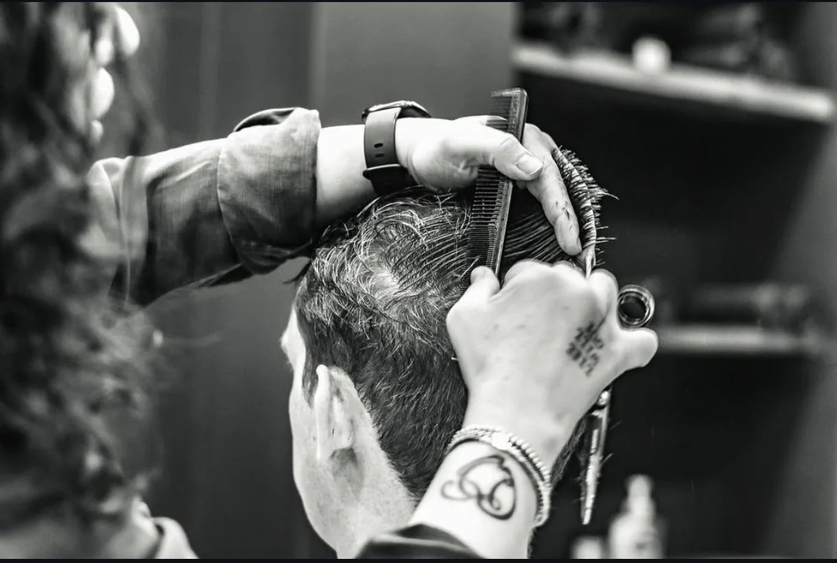 A hairstylist cutting a man's hair with scissors and using a comb, in a black and white photo.