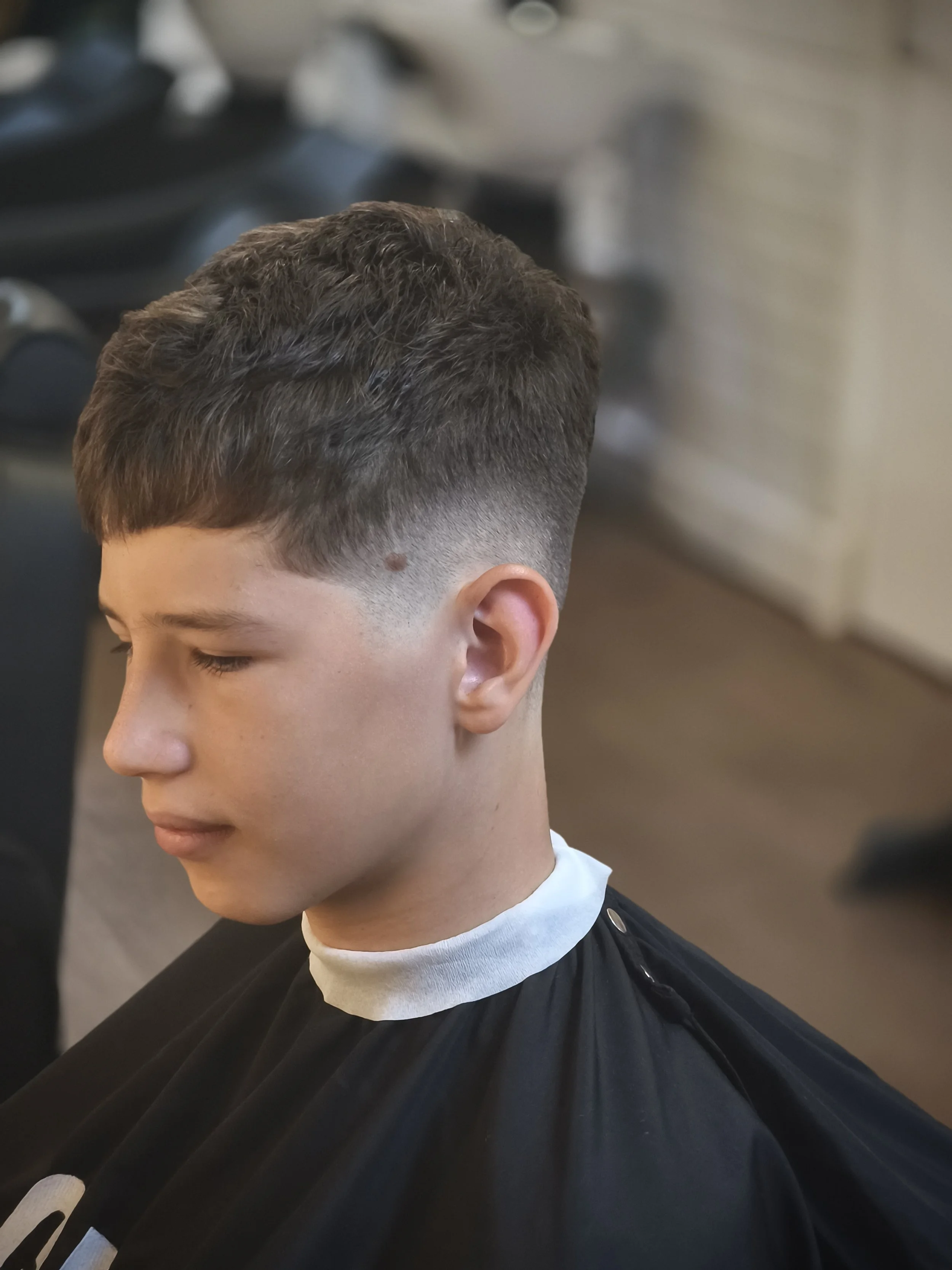 Young male with short, dark hair getting a haircut in a barber shop. He is sitting with a barber cape, and his hair is being styled or cut.