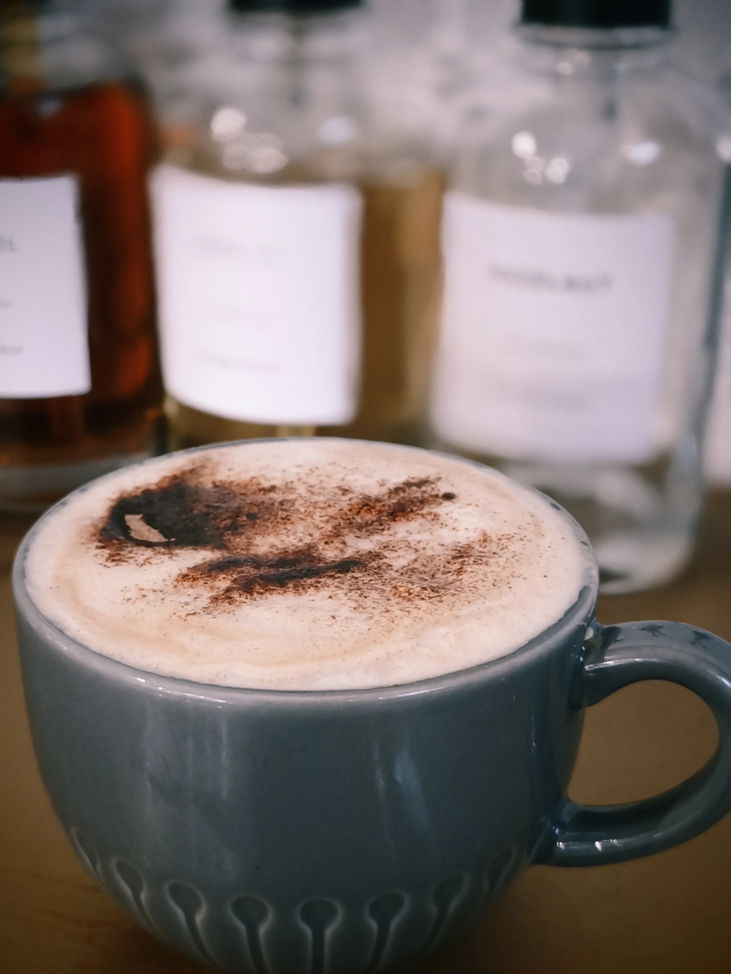 Close-up of a hot chocolate in a gray mug with decorative patterns, topped with foam and dusted with cocoa powder. In the background, there are bottles of syrup, one with a red cap and the other with a beige cap, slightly blurred.