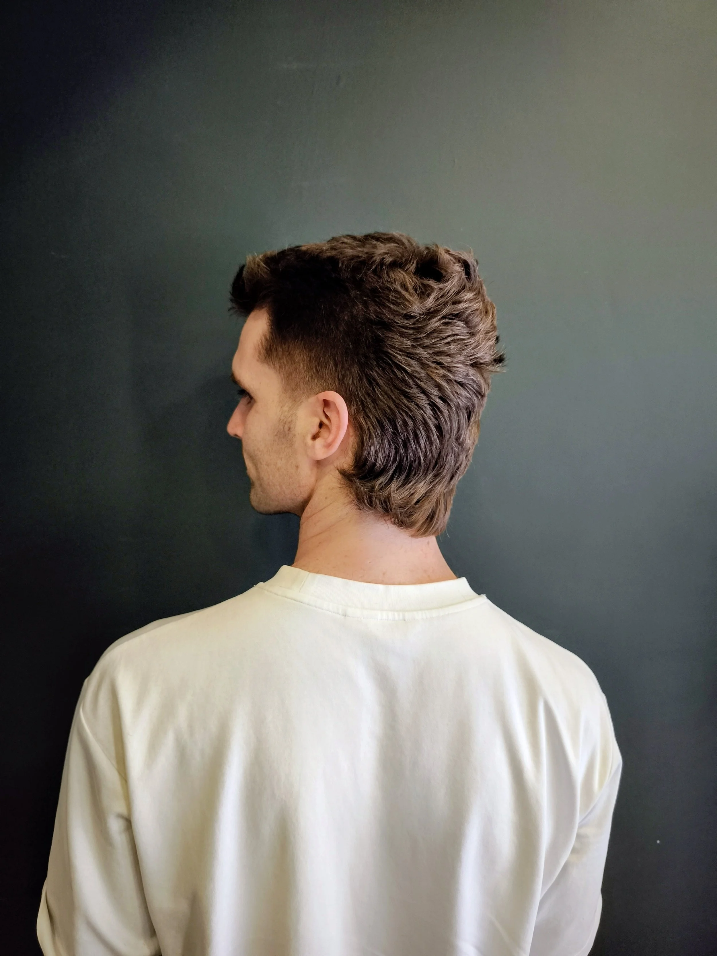Side view of a man with styled dark brown hair and a beard, wearing a white shirt, standing against a dark background.