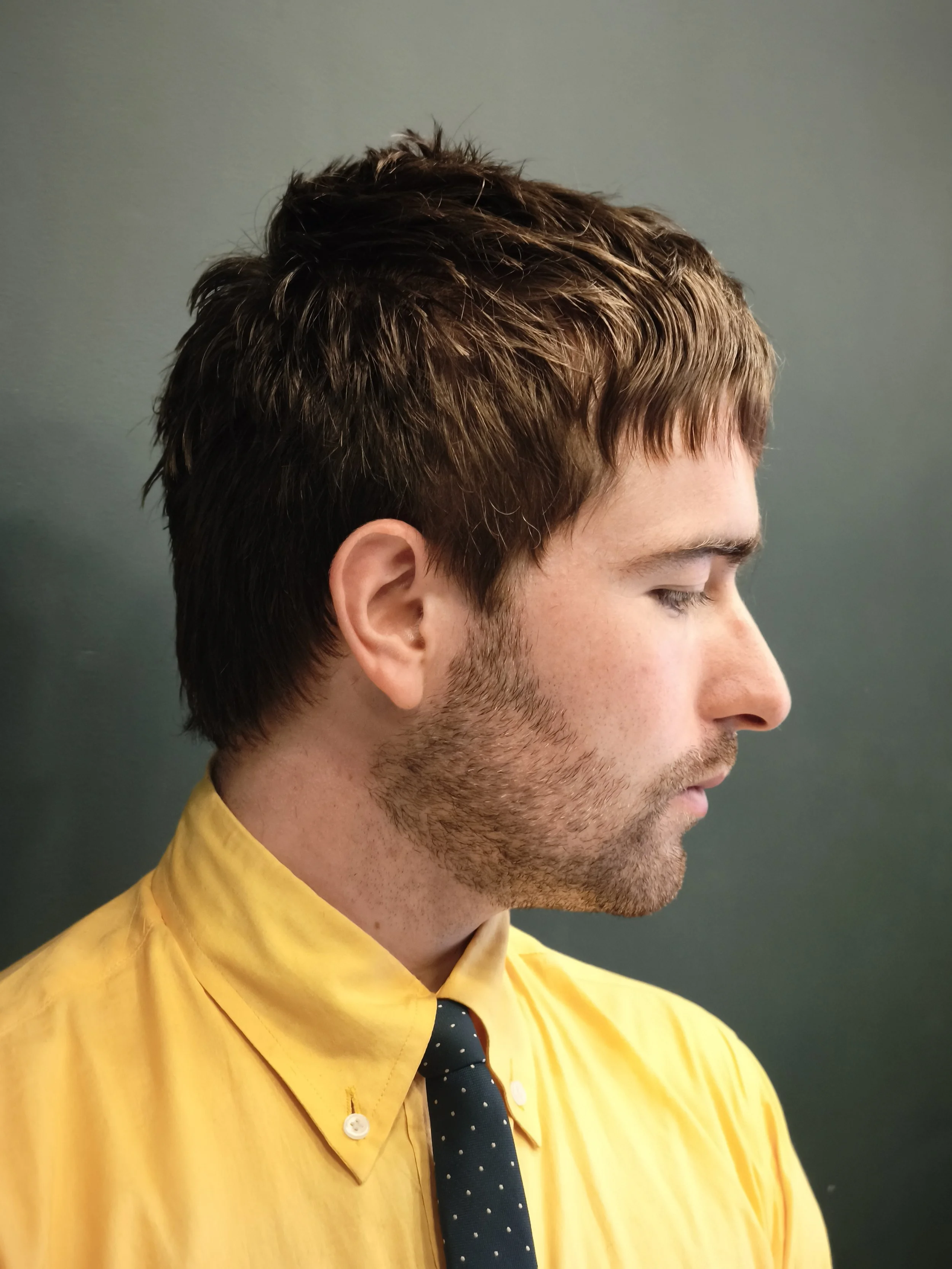 Side profile of a man with brown hair, wearing a yellow shirt and a dark blue polka dot tie, against a plain grey background.