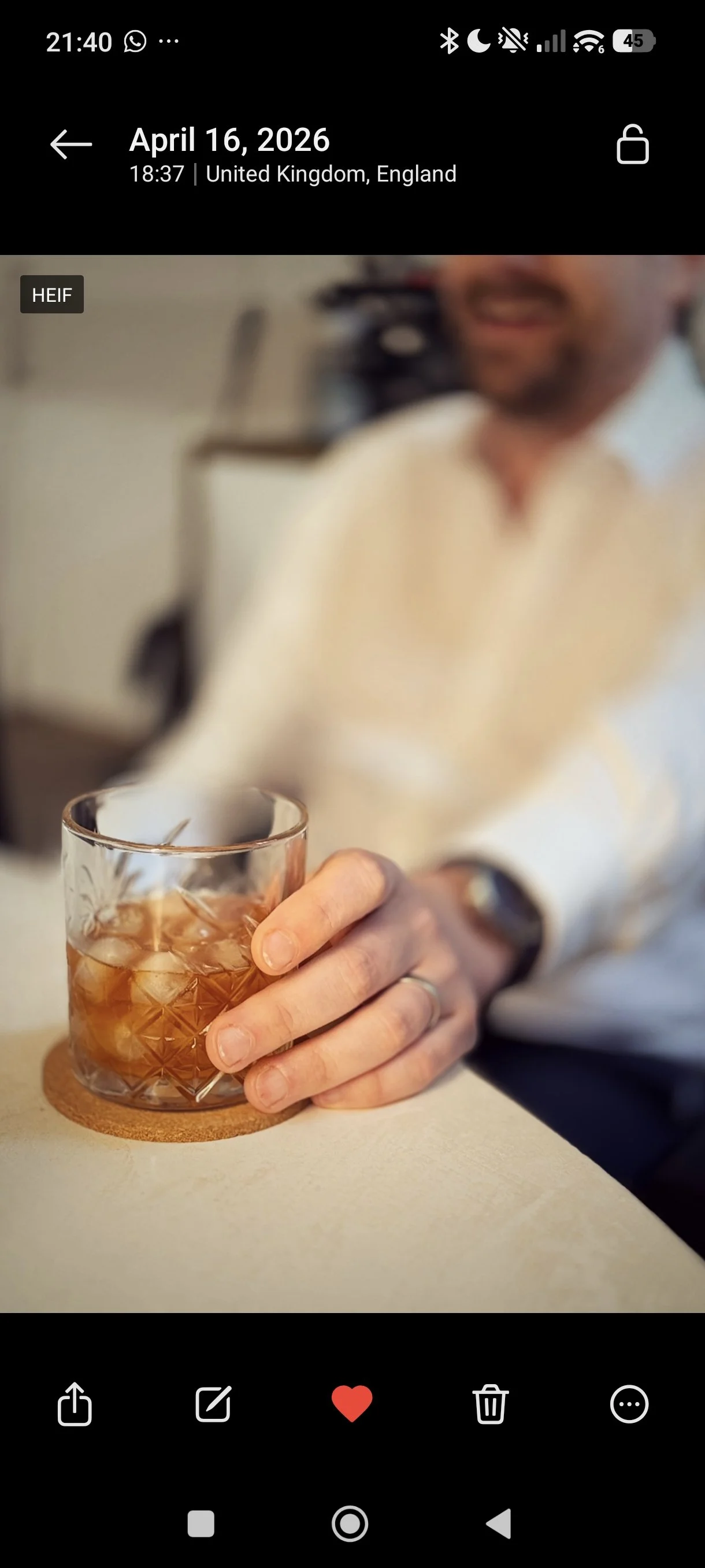 A person holding a glass of whiskey with ice, sitting at a table. The person's face is blurred, and they are wearing a watch and a ring on their finger.