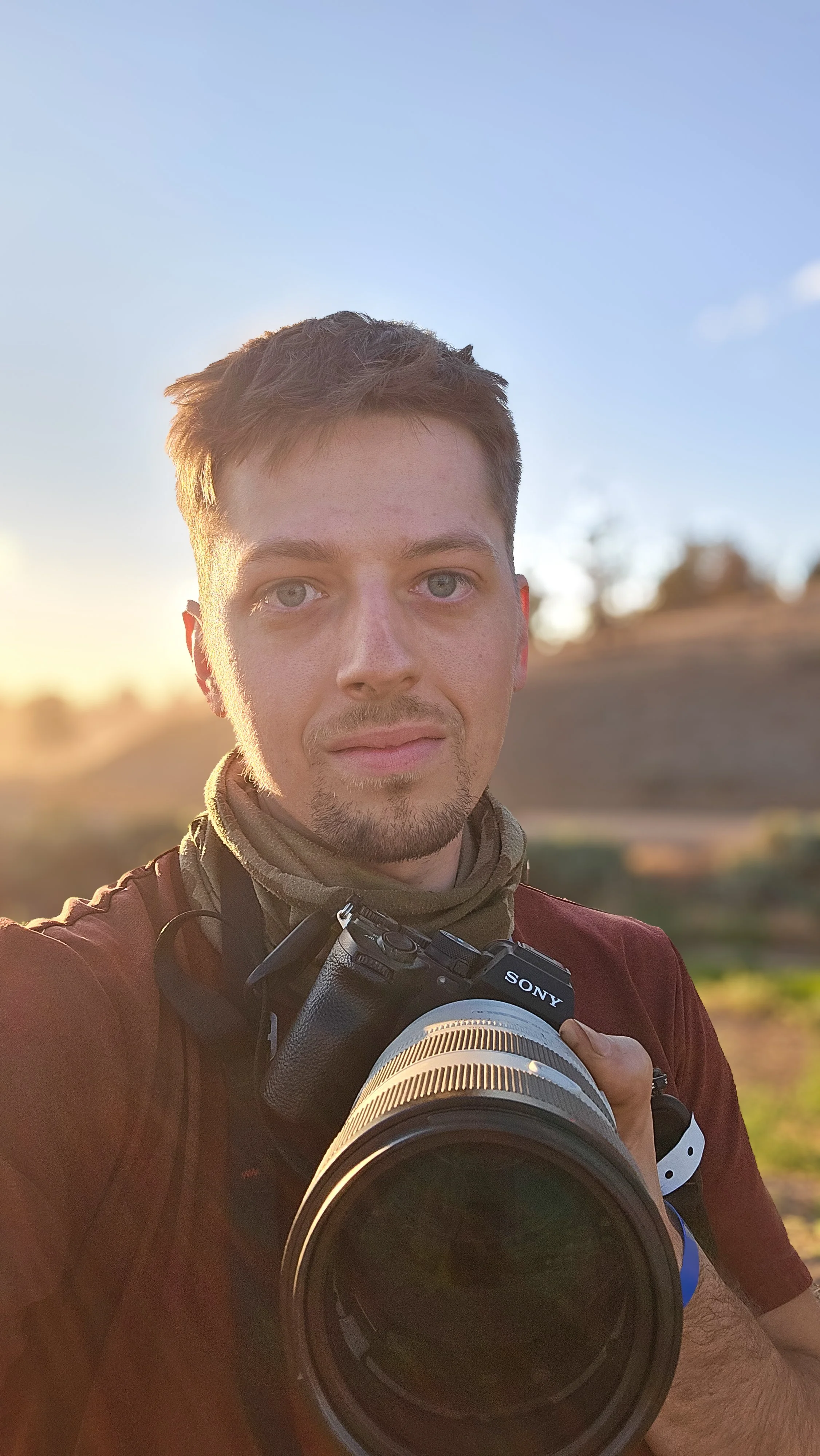 A young man with short brown hair and blue eyes holding a Sony camera outdoors during sunset, with a blurred landscape in the background.