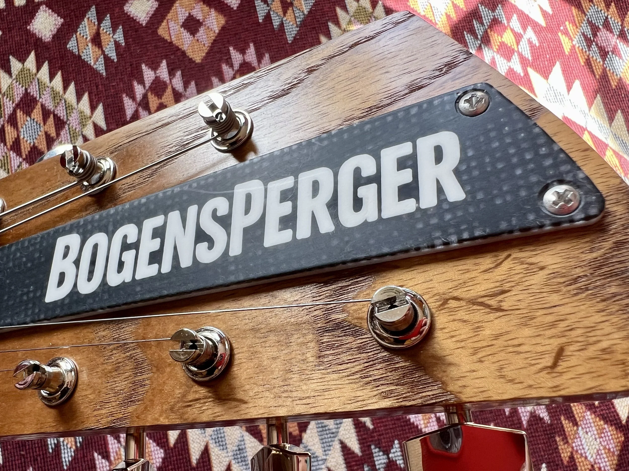 Ein Close-up von einem Gitarrenhals mit Stimmwirbeln und einem Schild, das 'BOGENSPEICHERG' aufgedruckt ist, auf einem bunten, geometrischen Teppich.