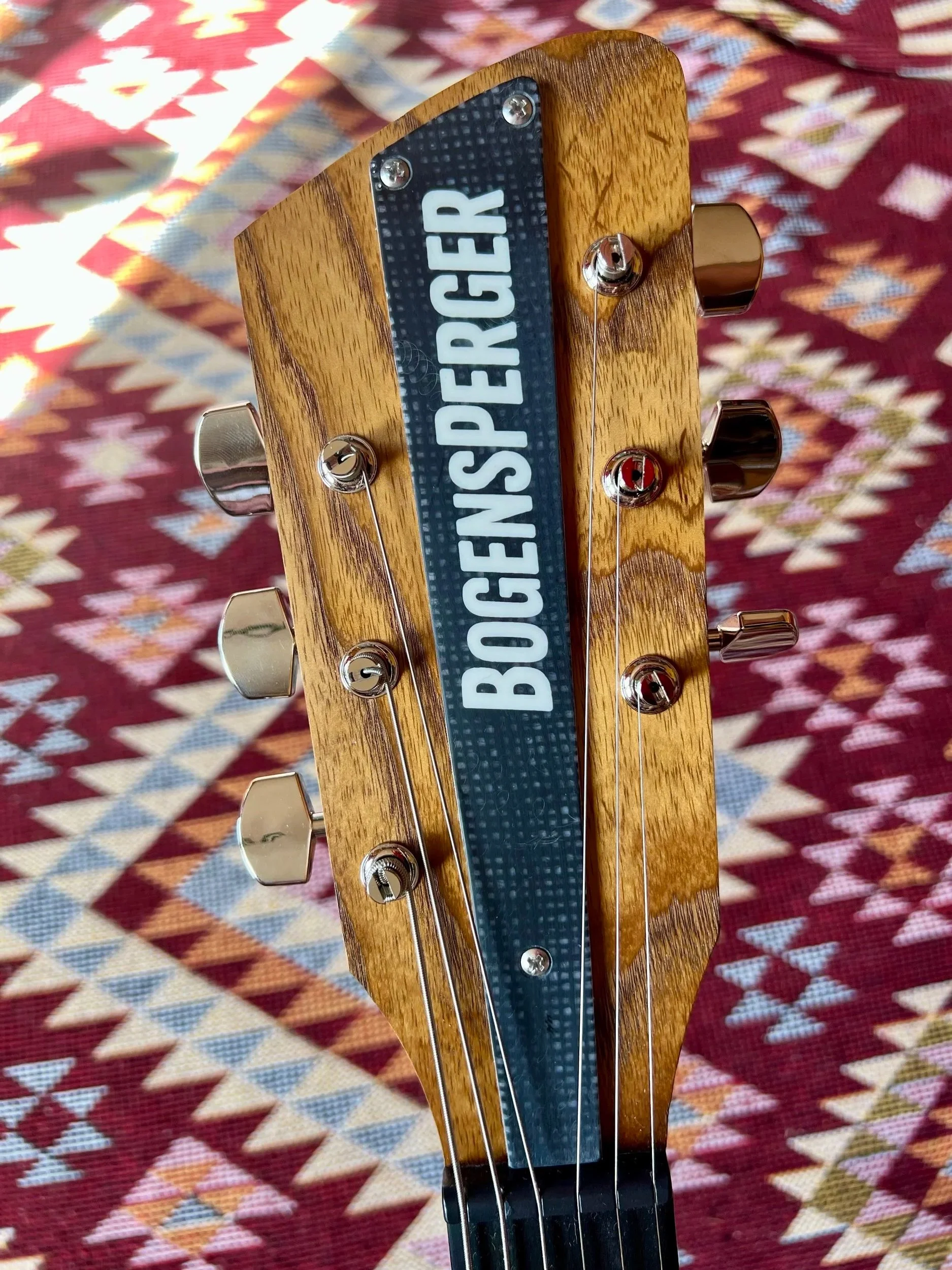 Der Kopf einer E-Gitarre mit einer Holzmaserung, einem schwarzen Gitarrenkopf mit weißen Buchstaben, die das Wort 'OBERSPENDER' bilden, und mehreren Stimmrollen, gegen einen bunten, geometrischen Stoffhintergrund.